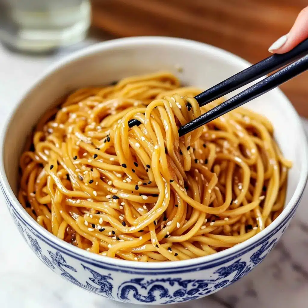 Hibachi Noodles, Japanese-style stir-fry noodles, quick hibachi stir-fry, easy homemade hibachi noodles, restaurant-style hibachi dish A white bowl with blue patterns on the outside is filled with glossy, golden-brown noodles mixed with black and white sesame seeds. The noodles appear thick and soft, loosely tangled and coated evenly with sauce. A woman's hand is holding black and natural wood chopsticks, lifting a small tangle of noodles from the bowl. The bowl sits on a wooden surface with a white and gray striped cloth beside it and blurred bottles in the background.