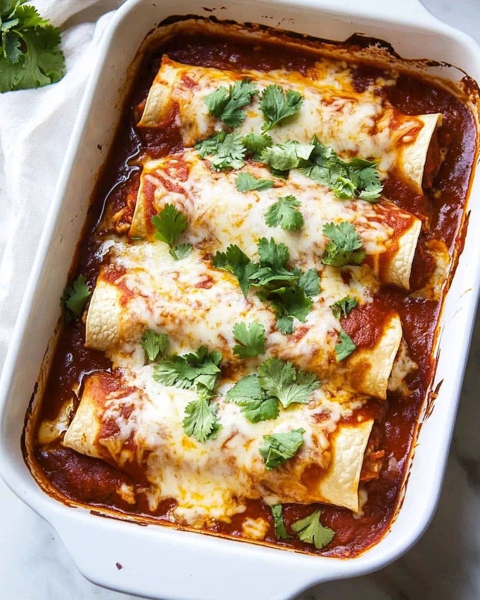 Beef Enchiladas, Beef Enchiladas Recipe, Homemade Beef Enchiladas, Easy Beef Enchiladas, Authentic Mexican Beef Enchiladas A white rectangular baking dish holds five rolled enchiladas arranged side by side, each covered in rich red sauce and melted white cheese. The cheese layer is thick and slightly browned on the edges, creating a creamy texture. Bright green cilantro leaves are scattered on top, adding fresh color contrast to the red sauce and white cheese. The edges of the tortillas peek through the sauce, showing their light golden color. The dish rests on a white marbled surface with a soft white cloth nearby.