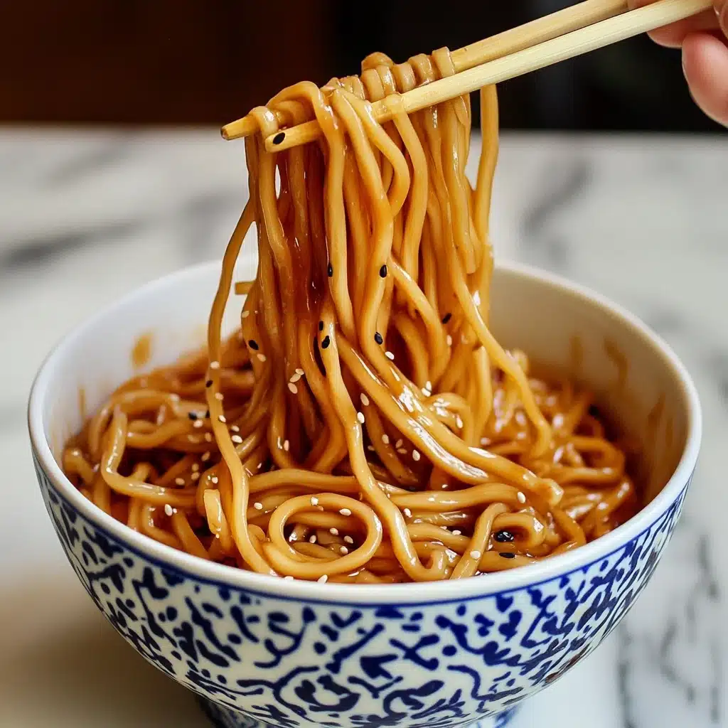 Hibachi Noodles, Japanese-style stir-fry noodles, quick hibachi stir-fry, easy homemade hibachi noodles, restaurant-style hibachi dish A bowl filled with cooked spaghetti noodles coated in a shiny brown sauce with black and white sesame seeds scattered throughout. The noodles are lifted mid-air by a pair of wooden chopsticks held by a woman's hand, showing their smooth and slightly glossy texture. The bowl is white with blue decorative patterns around its sides, sitting on a wooden surface with a white marbled texture in the background.