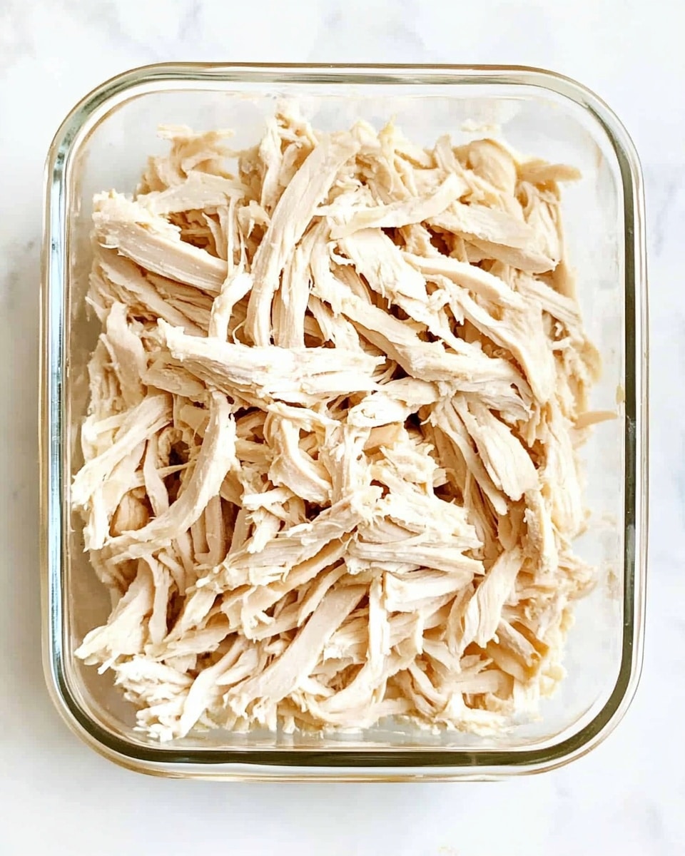 A clear rectangular glass container filled with shredded pale beige chicken pieces, arranged in loose layers that fill the container evenly from top to bottom, showing the fibrous texture of the meat strands closely packed together. The container is placed on a white marbled surface. photo taken with an iphone --ar 4:5 --v 6.1