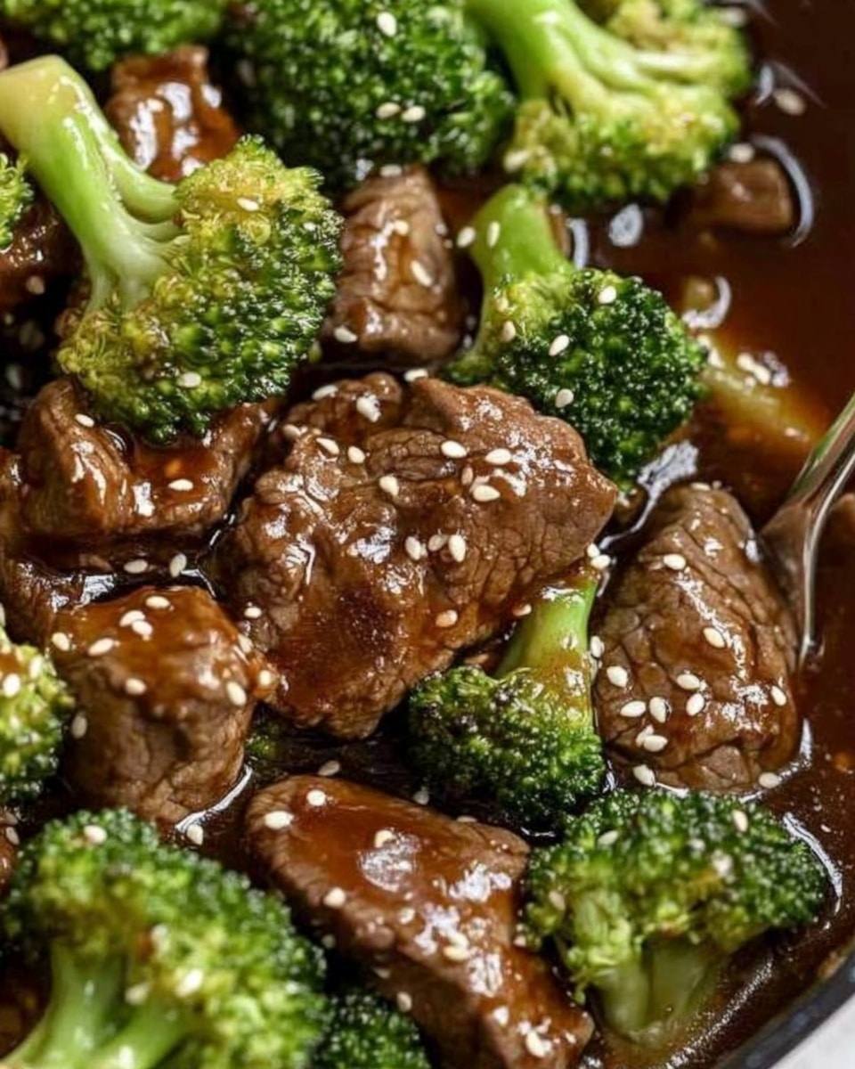 Chinese Beef and Broccoli Stir Fry, beef broccoli stir fry recipe, easy Chinese stir fry, quick beef and broccoli dish, savory Asian stir fry A white plate holds a bed of white rice as the bottom layer, soft and fluffy in texture. On top, there are bright green broccoli pieces with a fresh, slightly bumpy texture. Mixed with the broccoli are medium brown slices of beef with a glossy, juicy look from a thick brown sauce that coats both the meat and vegetables. The dish is sprinkled with small, round white sesame seeds for added texture and contrast. Two light brown wooden chopsticks rest on the edge of the plate. The plate is placed on a surface with a white marbled texture