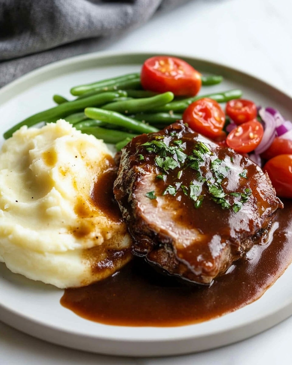 Oven Baked BBQ Chicken, homemade barbecue sauce, easy BBQ chicken recipe, baked chicken dinner, flavorful oven-baked chicken The image shows a close-up of a white plate with one thick layer of brown glazed meat on top, topped with small green herb pieces. Underneath the meat is a creamy, pale yellow mash that looks soft and smooth. Around the meat is a rich, glossy brown sauce pooling on the plate. In the blurred background, parts of halved red cherry tomatoes and green beans add bright red and green colors. A silver fork is on the right, holding a small piece of the meat. The plate sits on a white marbled surface.