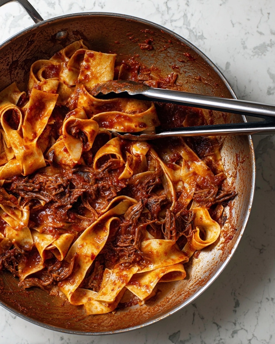 Inside a deep silver pan on a white marbled surface, wide flat pasta noodles are mixed with a rich, dark red tomato sauce that appears thick and chunky with pieces of cooked meat spread unevenly throughout. The pasta is tangled loosely among the sauce, showing a glossy texture with a soft, smooth look. A pair of metal tongs with black rubber handles lay inside the pan, resting over the pasta. The sauce clings to the sides of the pan, leaving a slightly oily residue. The setting is bright and clean. Photo taken with an iphone --ar 4:5 --v 7