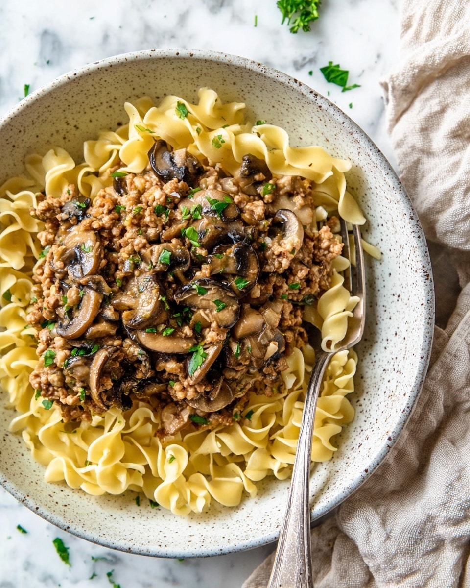 A white speckled bowl on a white marbled surface holds a dish with two main layers. The bottom layer is a bed of yellow egg noodles with a soft, slightly shiny texture, loosely piled in the bowl. On top is a creamy mushroom sauce mixed with small pieces of browned ground meat, light brown in color. Dark brown sautéed mushroom slices and translucent cooked onion pieces are scattered throughout the sauce. Bright green parsley leaves are sprinkled on top, adding a fresh pop of color. A silver fork rests in the bowl on the right side, with some noodles and sauce on it. A beige cloth is partially visible in the upper right corner. photo taken with an iphone --ar 4:5 --v 7
