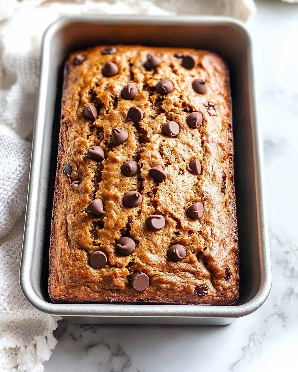 A top view of a single thick loaf of banana bread in a metal rectangular baking pan lined with parchment paper on two sides, the bread is golden brown with a rough textured surface and scattered dark brown chocolate chips spread unevenly on top, the background is a white marbled surface with part of a white cloth visible on the left side. photo taken with an iphone --ar 4:5 --v 6.1