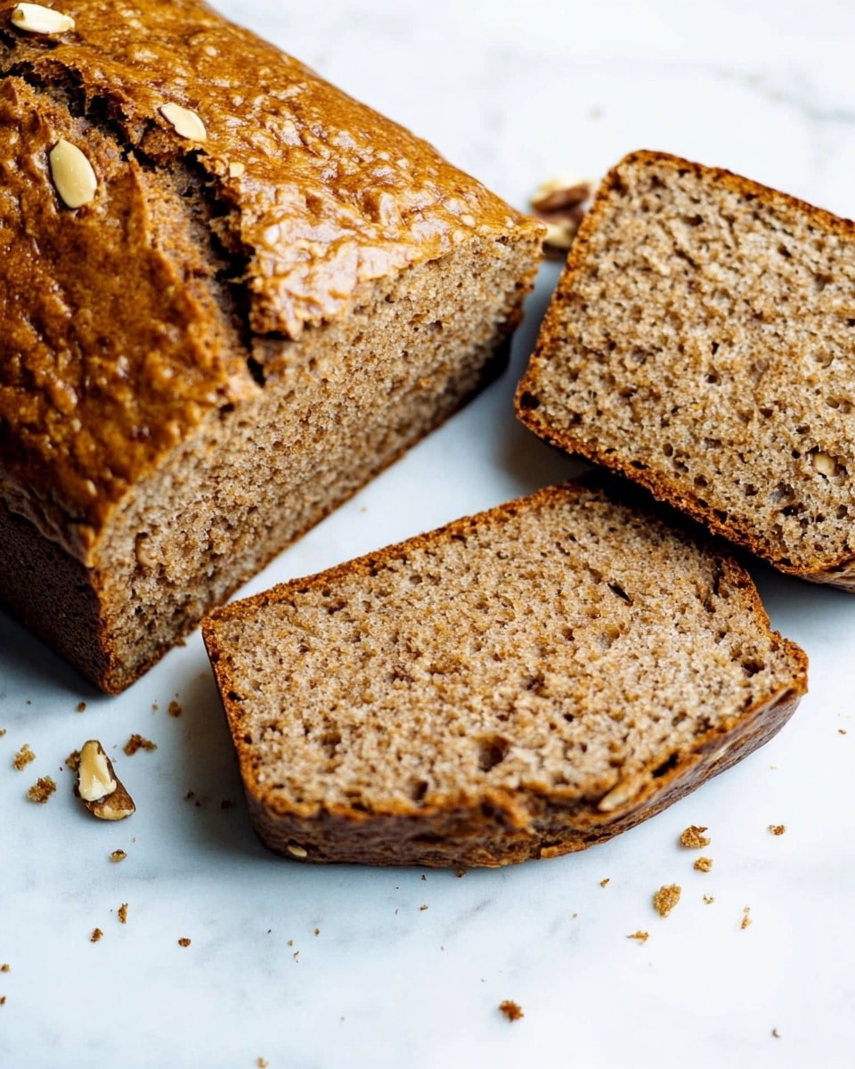 A golden-brown loaf of bread with a slightly cracked top is placed on a white marbled surface. The loaf is sliced from one end, showing a moist, dense inside speckled with small bits that look like nuts or seeds. Two slices lie flat next to the main loaf, revealing a consistent texture and color throughout. The background has a soft focus with a dark navy cloth in the corner. photo taken with an iphone --ar 4:5 --v 6.1