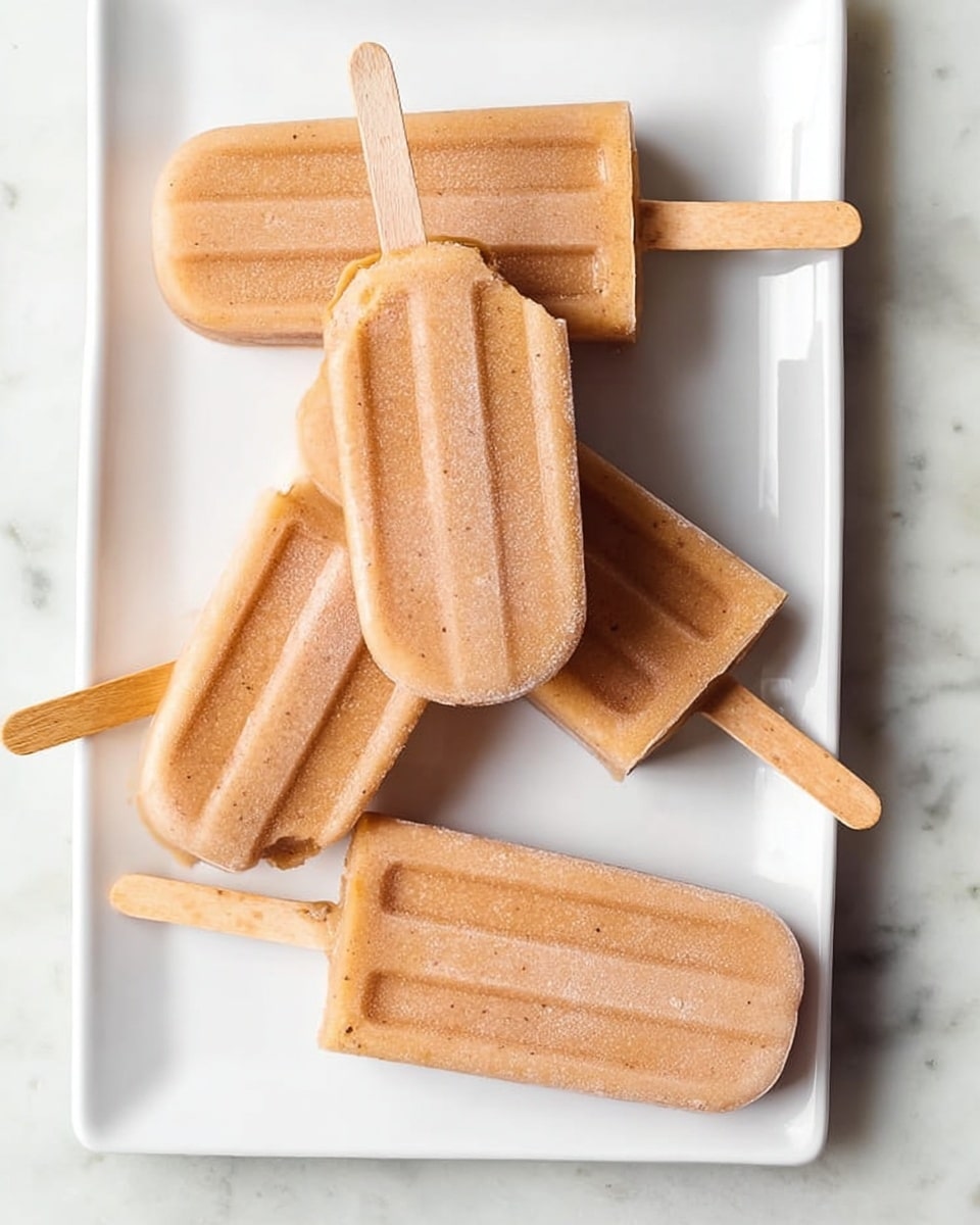 There are five light brown popsicles with small dark flecks on a white rectangular plate placed on a white marbled surface. Four popsicles lay flat on the plate, all showing smooth, even texture, and one popsicle is placed diagonally on top with a bite taken from the upper left corner, revealing a creamy orange inside. Each popsicle has a wooden stick extending outward. Photo taken with an iphone --ar 4:5 --v 7