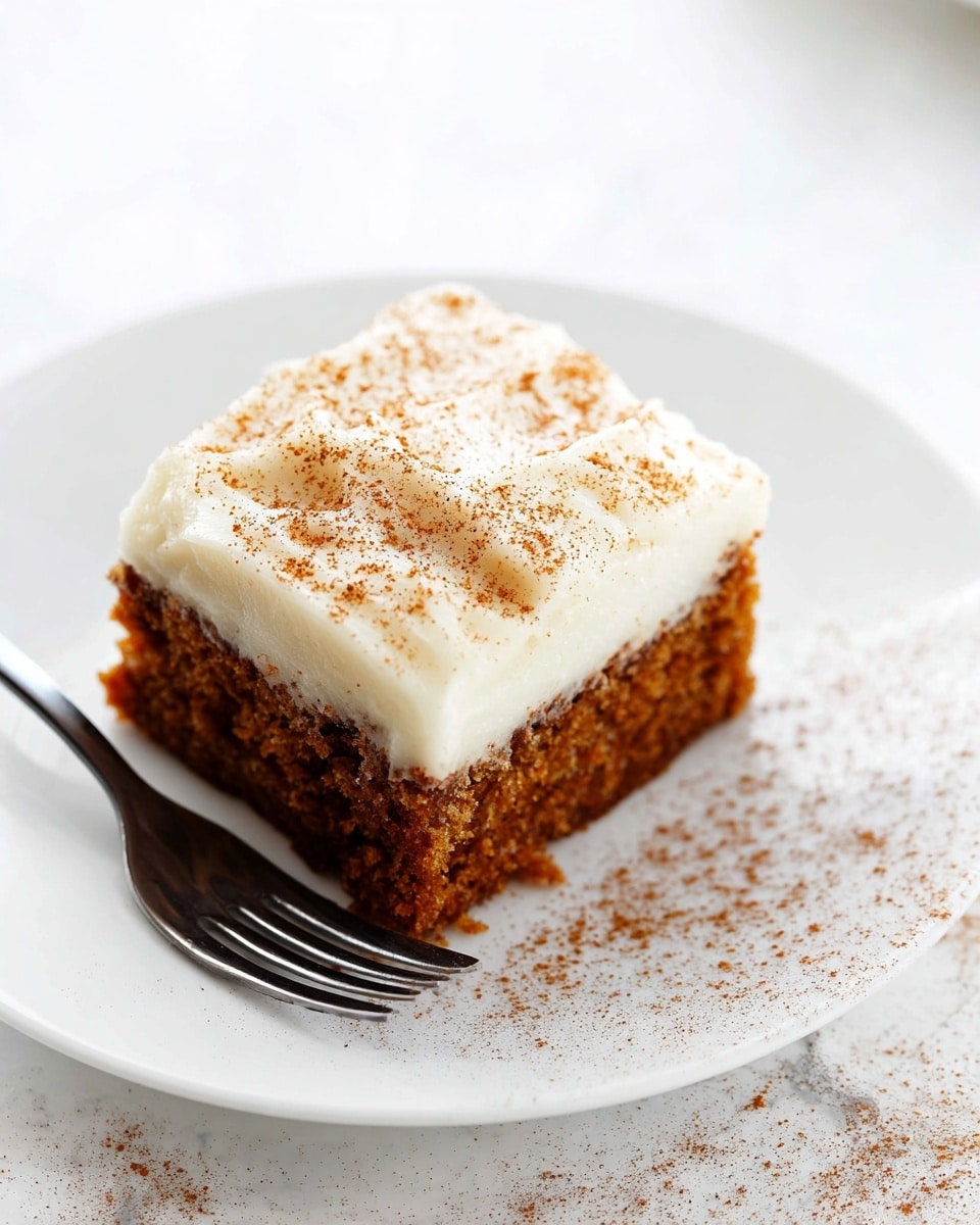 A square piece of carrot cake sits centered on a white plate, showing two layers: a thick, moist, deep orange-brown cake base with a soft, slightly crumbly texture, topped by a smooth, creamy layer of white frosting sprinkled lightly with fine brown cinnamon powder. A silver fork rests beside the cake slice at the edge of the plate. The plate is placed on a white marbled surface with soft natural light illuminating the scene, giving the dessert an inviting and fresh look. Photo taken with an iphone --ar 4:5 --v 7