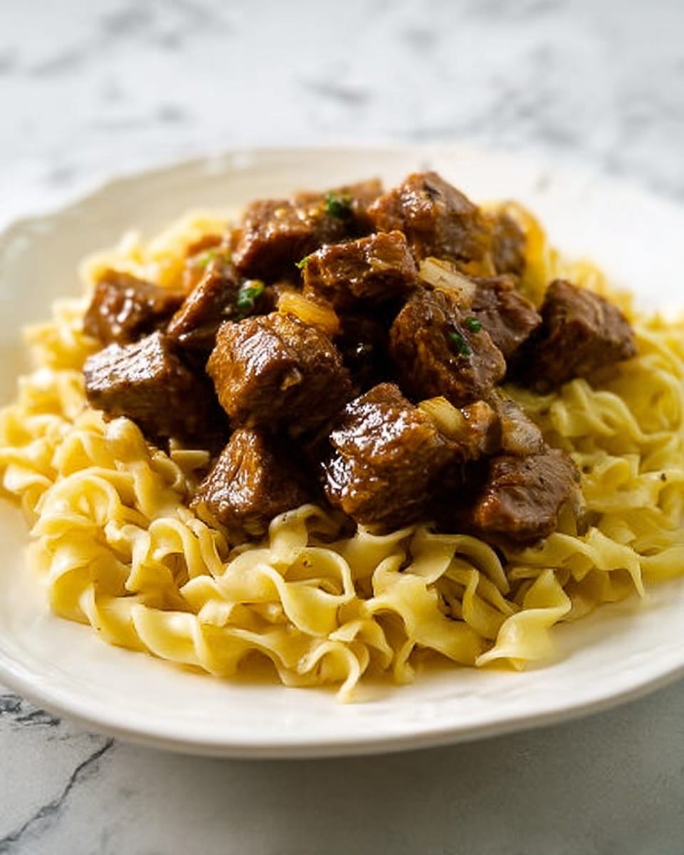 The image shows a white plate with two main layers of food. The bottom layer is a bed of light yellow egg noodles that are twisted and soft in texture. On top, there are chunks of dark brown stewed beef that look tender and juicy with a slight shine from the sauce, sprinkled with small bits of orange carrot and green herbs. The plate sits on a white marbled surface. Photo taken with an iphone --ar 4:5 --v 7