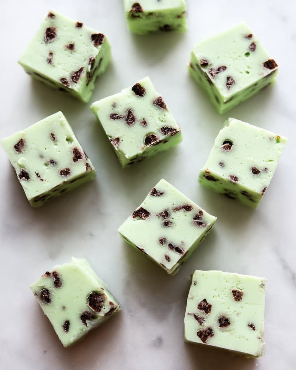 The image shows eleven small square pieces of light green fudge with dark brown chocolate chips spread throughout. Each piece is smooth on the top and has clean, straight edges. The fudge squares lie flat, scattered randomly on a white marbled surface. The minty green color is soft and pastel, contrasting with the irregularly placed tiny dark chocolate bits. The scene is well-lit with soft shadows giving depth to the fudge cubes. photo taken with an iphone --ar 4:5 --v 7