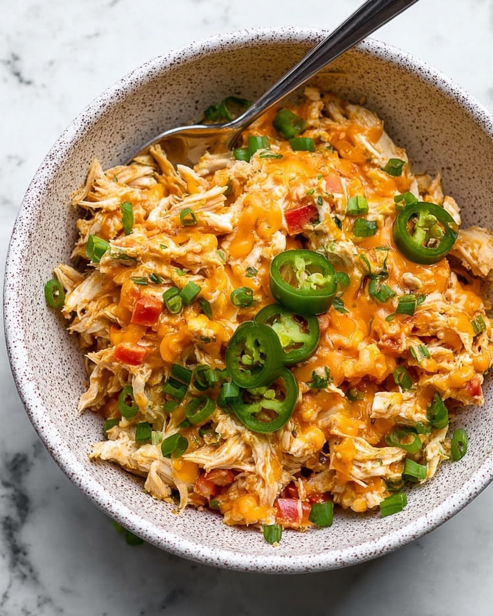 A white speckled bowl filled with shredded cooked chicken mixed with small diced red bell peppers and onions, topped with melted orange cheese sauce, thin slices of green jalapeño, and scattered green onion pieces. A silver spoon rests inside the bowl at the back, and green onion stalks are slightly visible in the top part of the frame. The background is a white marbled texture. Photo taken with an iphone --ar 4:5 --v 7