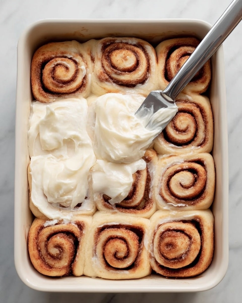 The image shows a white baking dish with twelve cinnamon rolls arranged in three rows of four. Each roll is light brown with darker brown swirls of cinnamon in the center, creating a spiral pattern on a soft, pale dough. On the top middle rolls, thick white frosting is being spread with a silver knife held by a woman's hand visible on the bottom right. The dish is set on a white marbled surface with a folded gray cloth partially visible at the bottom left. Photo taken with an iphone --ar 4:5 --v 7