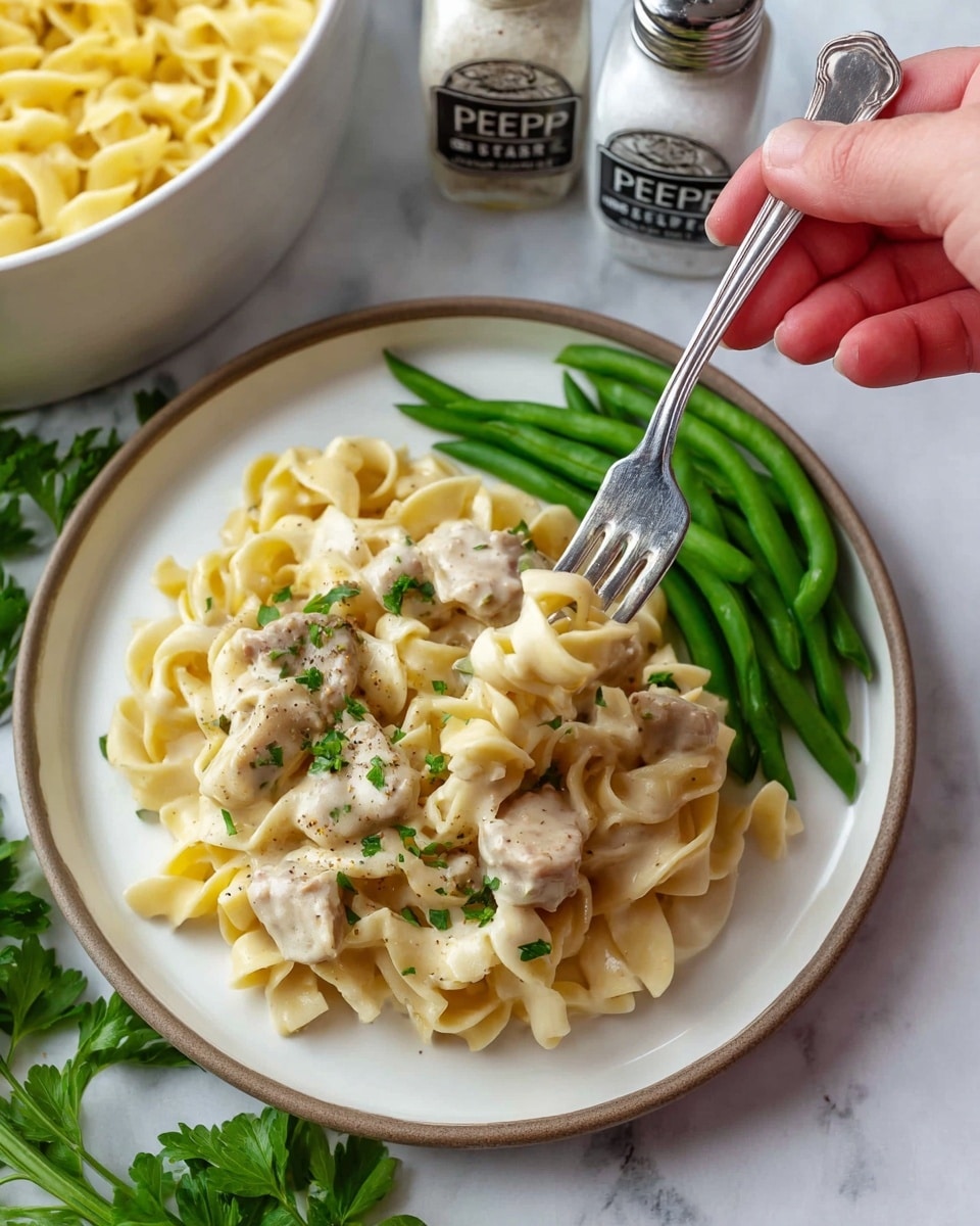 The image shows a white plate with two main layers of food. The bottom layer is a bed of light yellow egg noodles with a soft, curly texture. On top of the noodles are pieces of creamy white chicken with specks of black pepper and small green herb bits scattered over them. To the right of the plate is a neat pile of bright green beans, cooked but still crisp. A woman's hand holds a silver fork picking up a small cluster of noodles and chicken. The plate sits on a white marbled surface with some fresh green parsley leaves visible at the bottom edge, and there are some white salt and pepper shakers blurred in the background. photo taken with an iphone --ar 4:5 --v 7