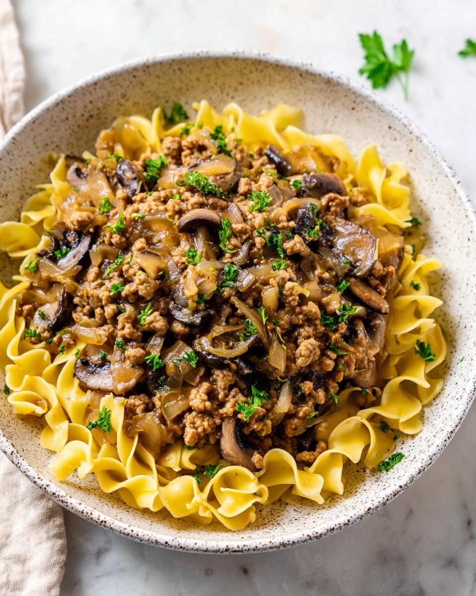A white speckled bowl filled with a base layer of pale yellow curly egg noodles. On top, there is a thick layer of cooked light brown ground meat mixed with cooked translucent onion pieces and dark brown sliced mushrooms. The dish is garnished with small sprinkles of bright green chopped herbs. The bowl sits on a white marbled surface with a few fresh green herb leaves scattered around. photo taken with an iphone --ar 4:5 --v 7