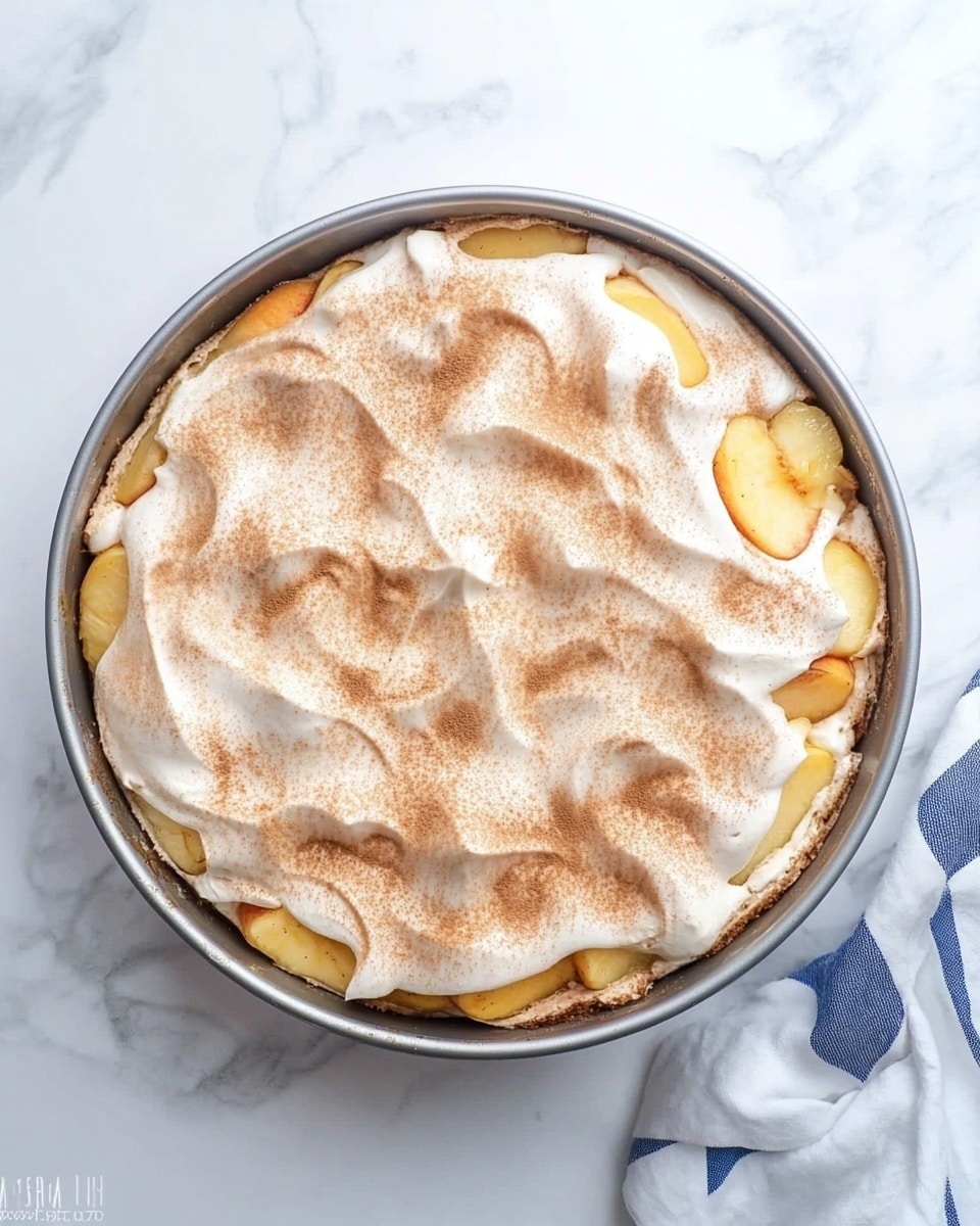 A round baking pan filled with two layers, the bottom layer is made of chunky yellow fruit pieces, visible around the edges. On top, a thick white creamy layer is spread unevenly, dusted with a light brown powder, likely cinnamon, giving a textured look with swirls and peaks. The pan sits on a white marbled surface next to a white and blue checkered cloth. photo taken with an iphone --ar 4:5 --v 6.1
