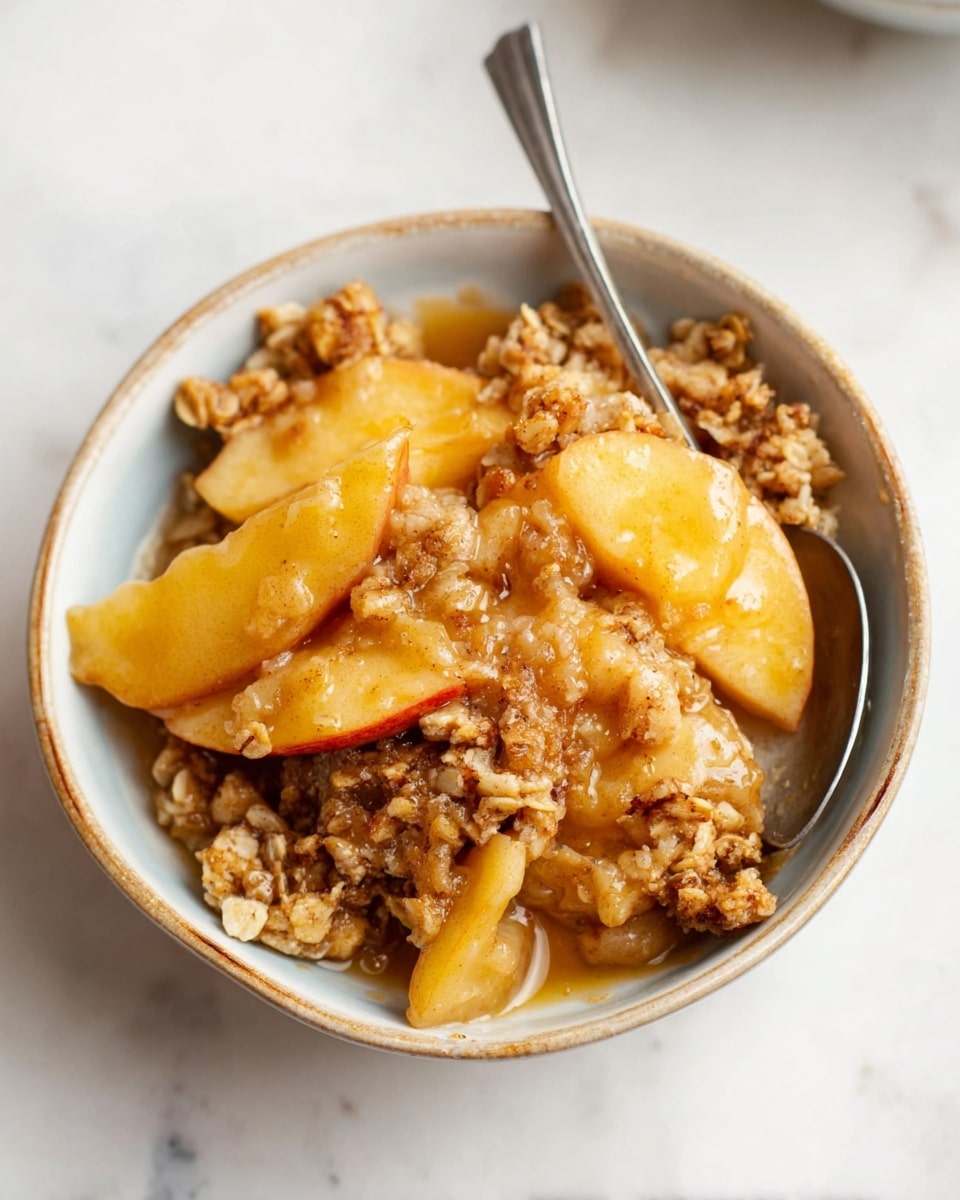 A close-up image of a bowl filled with a warm apple crisp dessert. The dish has visible layers starting with soft, golden-brown apple slices at the bottom, peeking through a thick, crumbly topping made of oats and brown sugar that looks crunchy and textured. The top layer has small clusters of crisp oats mixed with bits of apple, showing a mix of light beige and golden tones. A silver spoon is partially visible inside the bowl, resting against the side. The bowl is white and sits on a white marbled surface that is clean with a few stray oat crumbs nearby. Photo taken with an iphone --ar 4:5 --v 7