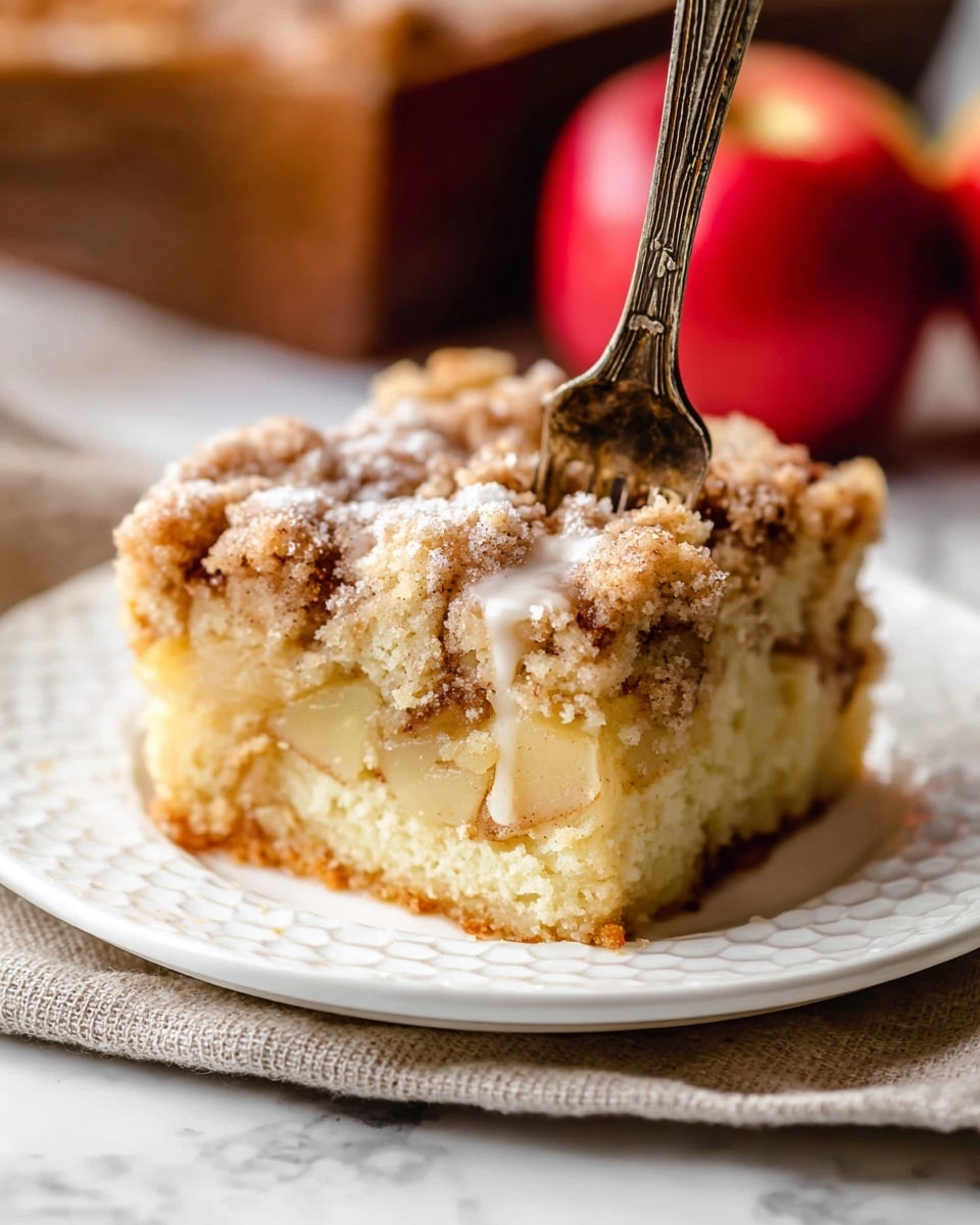 A thick slice of crumb cake sits at the center of a white plate, showing three visible layers: a crumbly, light brown topping with a rough texture; a soft, pale yellow cake middle layer scattered with small bits of apple; and a dense, slightly darker base layer. A vintage-looking fork with a rustic brown handle stands upright, its prongs digging lightly into the cake. The plate rests on top of another white plate with a honeycomb pattern, placed on a soft beige cloth that features faint circular designs. In the blurred background, there are two red apples adding a splash of color. The whole scene is set on a white marbled surface. Photo taken with an iphone --ar 4:5 --v 7