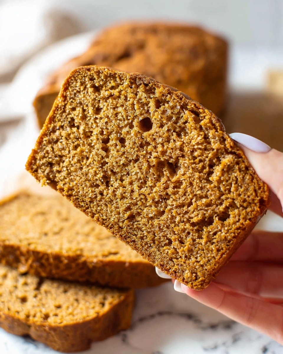 A close-up of a single slice of orange-brown bread being held by a woman's hand with pale pink nails, showing a moist and slightly crumbly texture with small holes inside. Behind the slice, there is a white loaf and several more slices stacked on a white marbled surface. The bread has a rough crust edge and an uneven surface, indicating a homemade rustic look. Photo taken with an iphone --ar 4:5 --v 7