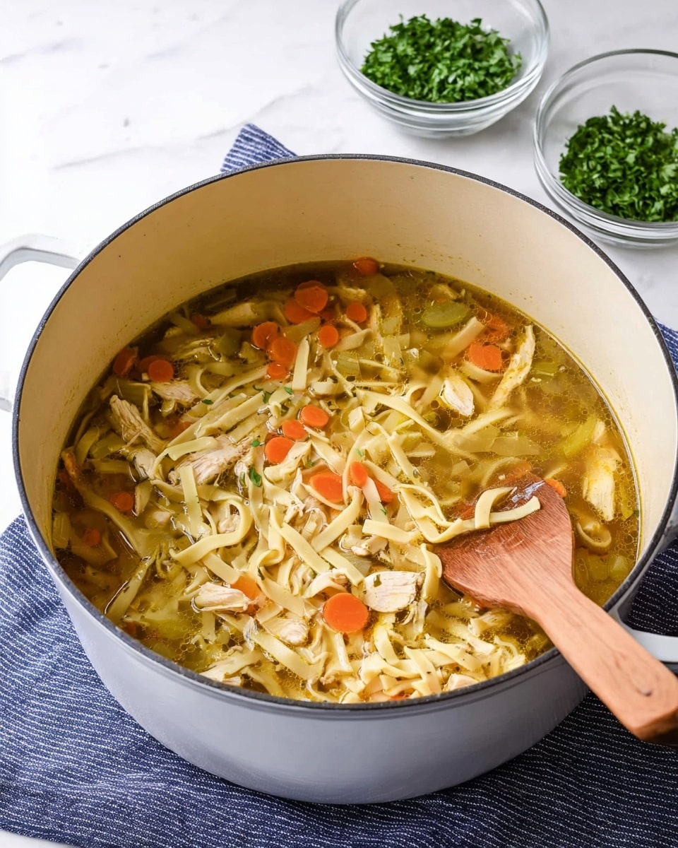 A large white pot sits on a white marbled surface, filled with a light yellow broth containing wide, flat noodles, pieces of light brown meat, small orange carrot slices, and bits of green herbs scattered throughout. A wooden spoon with a smooth, warm brown handle is partially submerged in the soup, resting against the side of the pot. In the background, there are pieces of torn bread and some green parsley, with a stack of white bowls stacked on top of each other. The scene has a cozy and fresh feel, photo taken with an iphone --ar 4:5 --v 7