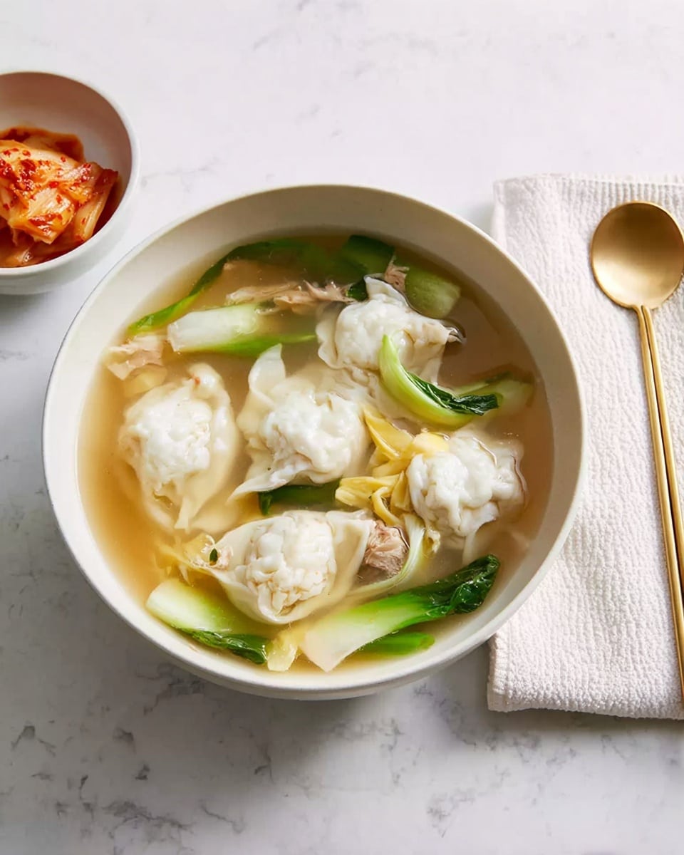 A white bowl filled with clear broth soup containing several white dumplings that have a soft, slightly wrinkled texture. The soup has light yellow egg ribbons and green slices of spring onion floating on top, along with small pieces of meat. Next to the bowl, there is a smaller white bowl filled with reddish-orange kimchi. On the right side, a white cloth napkin is placed underneath a golden spoon and golden chopsticks, resting on a white marbled surface. Photo taken with an iphone --ar 4:5 --v 7