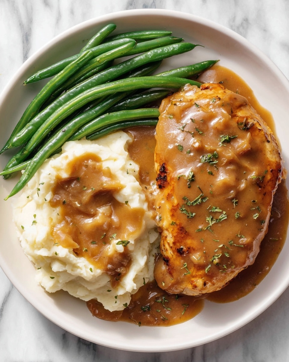 A white round plate holds a meal with three main parts: on the left are bright green cooked green beans, smooth and shiny with a fresh look; next to them on the top left is a golden-brown cooked chicken piece covered in a thick, rich brown gravy with visible small bits and green herb specks scattered on top; to the right, there is a large serving of creamy white mashed potatoes with a heavy layer of the same brown gravy poured over it, creating a textured, glossy surface with bits of onions or similar elements visible; the plate sits on a white marbled surface. photo taken with an iphone --ar 4:5 --v 7