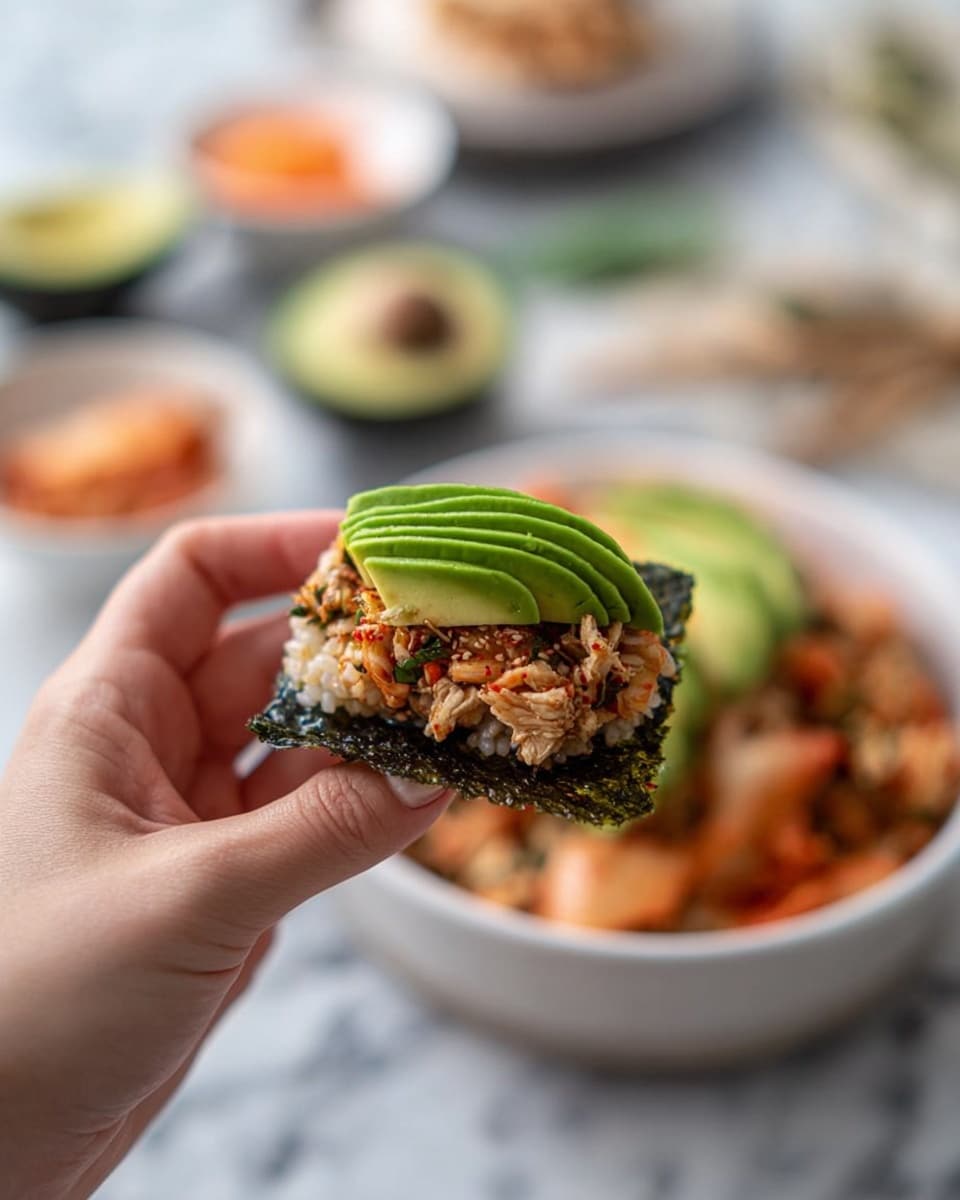 A white bowl filled with a rice dish mixed with small pinkish-orange pieces of cooked salmon, sprinkled with dark green seaweed flakes on top, arranged with five thin slices of green avocado fanned out on one side of the bowl; the bowl sits on a white marbled surface surrounded by a white plate with a piece of cooked salmon, a small white bowl with a spoon and light brown flakes, a rectangular white plate holding three sheets of dark green seaweed, and a small white dish filled with bite-sized orange pieces of kimchi; a green spice grinder is partly visible in the background, and a crumpled gray cloth napkin is placed near the bottom left corner photo taken with an iphone --ar 4:5 --v 7