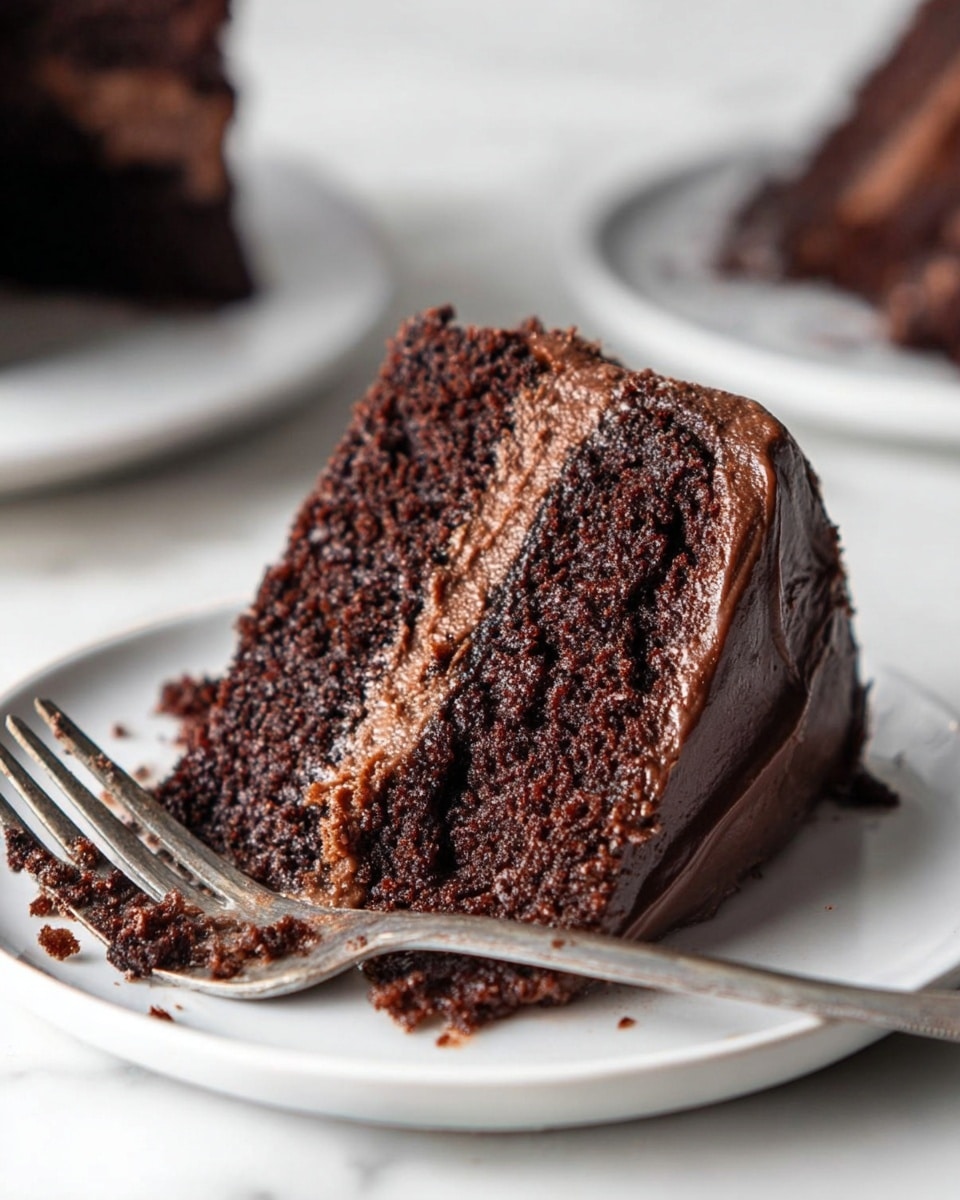 A close-up of a thick slice of three-layer dark chocolate cake on a white plate with a shiny silver fork resting beside it. Each cake layer is rich and moist with a slightly crumbly texture. Between the layers and covering the top and sides is smooth, creamy chocolate frosting. The plate sits on a white marbled surface with soft natural light highlighting the cake’s dark, rich brown color and the glossy finish of the frosting. Photo taken with an iphone --ar 4:5 --v 7