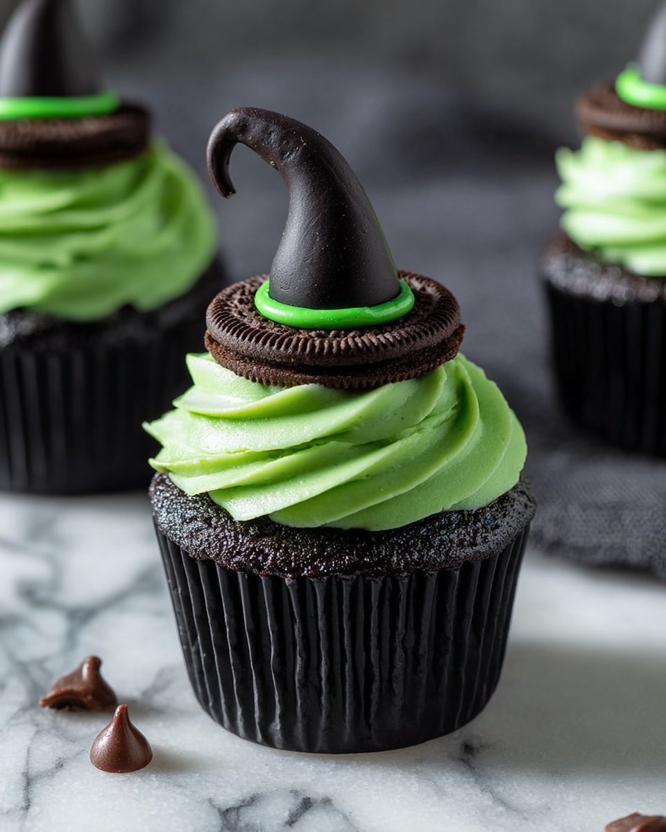 The image shows a close-up of a cupcake with three main layers. The bottom layer is a dark gray cake with a soft texture inside a black paper wrap. On top of the cake is a swirl of bright green smooth frosting. Sitting on the green frosting is a dark brown cookie ring, and on top of the cookie is a tall, black witch hat made of a smooth, slightly shiny material with a green band at the base. The cupcakes are placed on a white marbled surface with a few scattered chocolate chips nearby. Photo taken with an iphone --ar 4:5 --v 7