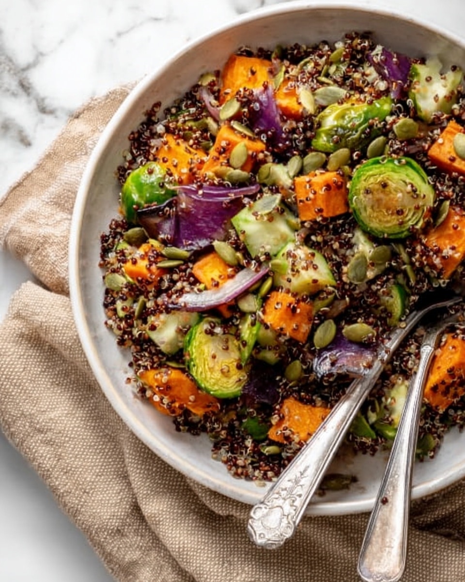 The image shows a clear glass bowl filled with a colorful fall quinoa salad. Inside the bowl, there are big chunks of bright orange roasted squash, deep purple onion pieces, small green Brussels sprouts, and dark red quinoa mixed with fresh green spinach leaves. The bowl rests on a white marbled surface, next to a small glass jar with a mustard yellow dressing inside. A white cloth is placed nearby, and a red lid with a white label reading