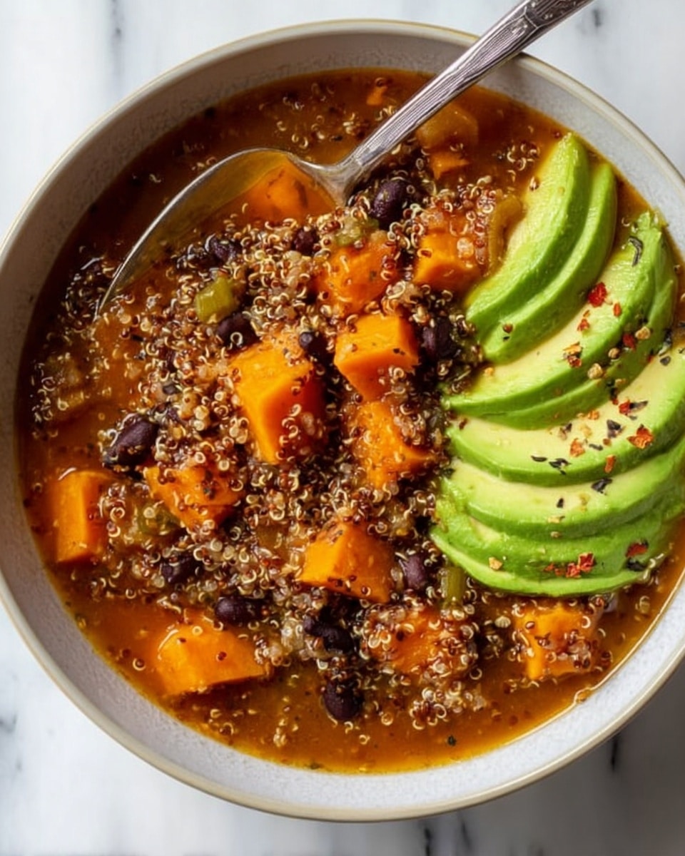 The image shows a white bowl filled with a warm quinoa stew made of small, light brown quinoa grains mixed with black beans and chunks of orange sweet potato in a reddish broth. On the right side of the bowl, there is a neatly sliced bright green avocado, slightly fanned out. A silver spoon is placed inside the bowl on the left side, partially submerged in the stew. The bowl sits on a white marbled surface, adding a clean and fresh background to the dish. photo taken with an iphone --ar 4:5 --v 7