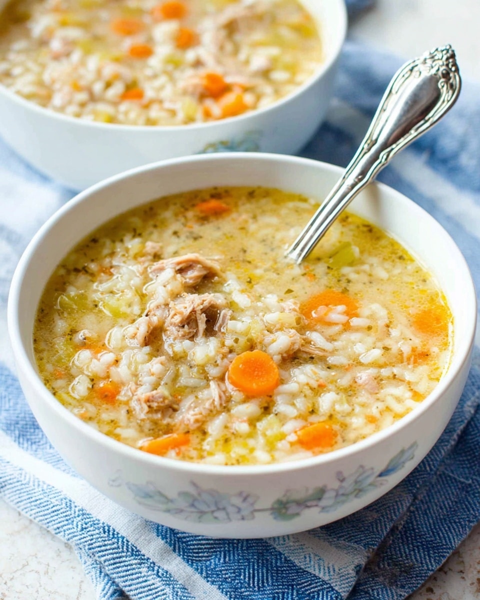 Two white cups with gray floral designs are filled with thick chicken and rice soup. The soup has visible layers of light broth, white cooked rice, small orange carrot pieces, and shredded light brown chicken chunks mixed with green celery bits. One cup is full and has a silver spoon inside, showing the texture of the soup clearly. The cups sit on a light blue and white striped cloth with a shiny silver spoon nearby on the white marbled surface. Photo taken with an iphone --ar 4:5 --v 7