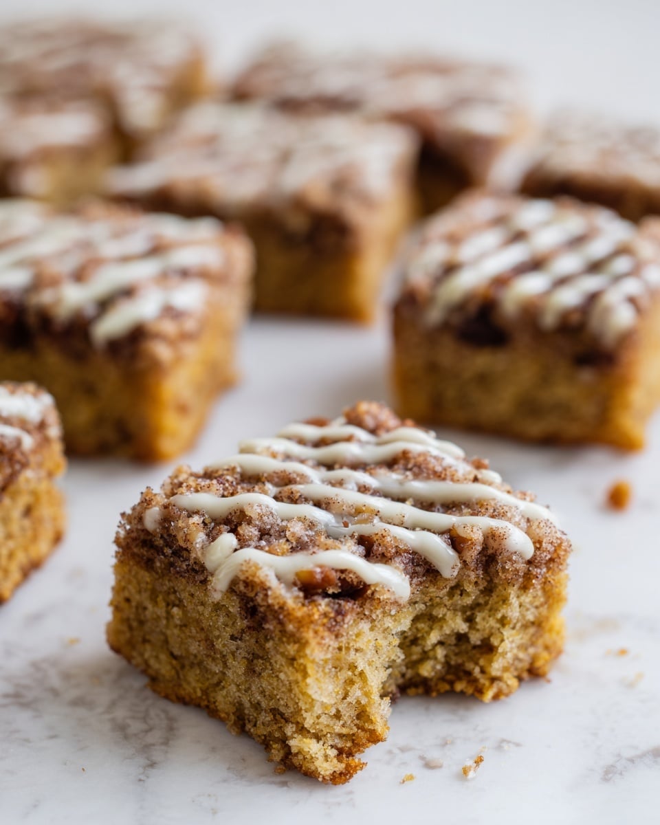 The image shows nine squares of a light brown cake with a soft texture, topped with a crumbly layer mixed with small dark brown pieces and drizzled with a light-colored glaze. One square is placed in the front right corner and has a bite taken out of it, revealing a moist, crumbly inside with small dark bits scattered throughout. The other eight squares are neatly arranged in rows in the background on a white marbled surface. The cake squares have crumbly tops with rough textures and lighter glaze lines on top, creating contrast in color and texture. Photo taken with an iphone --ar 4:5 --v 7