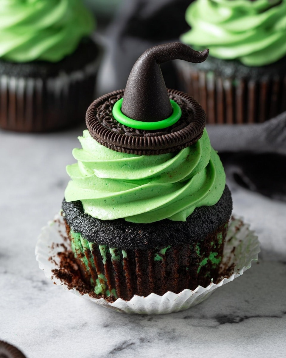 This image shows a dark chocolate cupcake with chocolate chips inside, wrapped in a white paper liner partially peeled down. It has one thick layer of bright green frosting swirled on top. On the frosting, a whole chocolate sandwich cookie sits flat, and on top of the cookie is a small black witch hat made of a textured material, with a green band at its base. The background is a white marbled texture. photo taken with an iphone --ar 4:5 --v 7