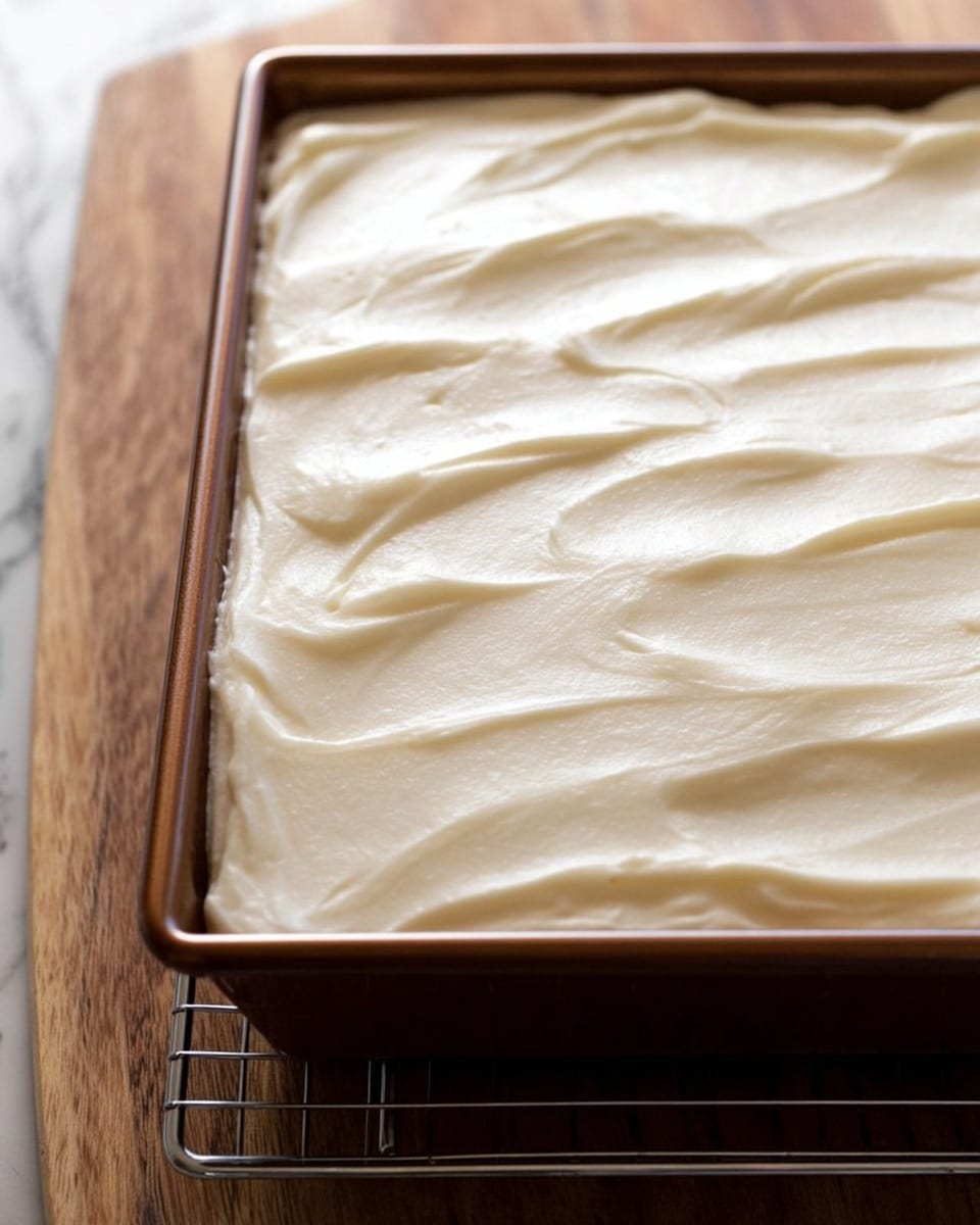 A single piece of moist carrot cake with a rough texture and visible small orange carrot bits is placed on a white plate. The cake has one thick layer of creamy white frosting on top, spread smoothly but with slight waves. Next to the cake is a shiny silver fork resting partly on the plate. In the background, there is a white baking dish with the rest of the carrot cake, which also has the thick white frosting layer evenly spread on top, with one missing piece. The surface beneath is a light wood texture, and a green cloth with white and red flower patterns is partly visible near the plate. Photo taken with an iphone --ar 4:5 --v 7