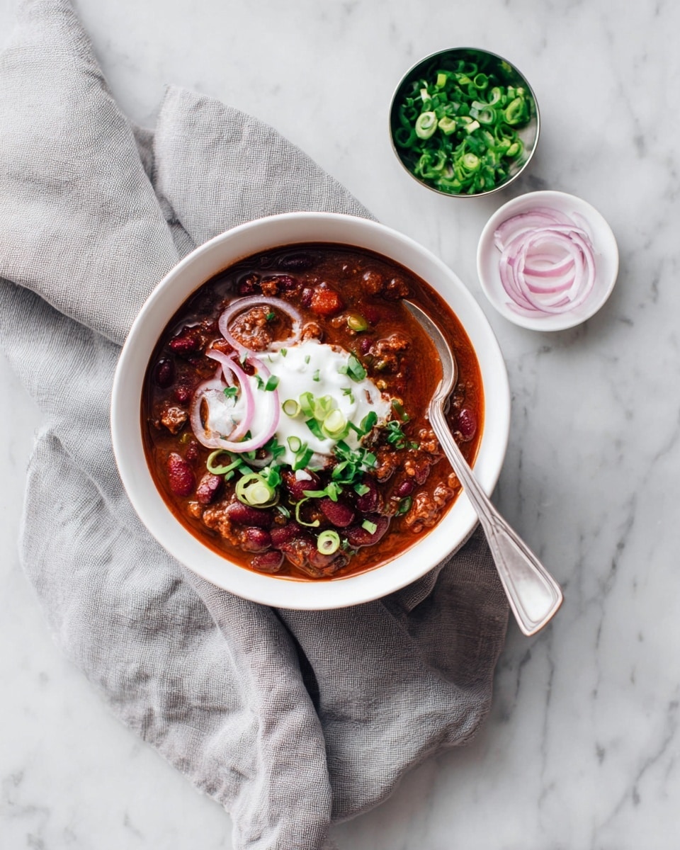 A white bowl filled with dark red chili that has visible kidney beans, black beans, ground meat, and tomato chunks forming the base layer. On top, there is a dollop of white sour cream with thin light purple onion rings and green chopped spring onions scattered over it. A silver spoon rests inside the bowl, with its handle pointing outward. The bowl sits on a soft gray cloth, placed on a white marbled surface. Nearby, there is a small white dish with thin light purple onion rings and a small metal bowl filled with chopped green spring onions. A few pieces of green chopped spring onions are scattered on the surface too. photo taken with an iphone --ar 4:5 --v 7