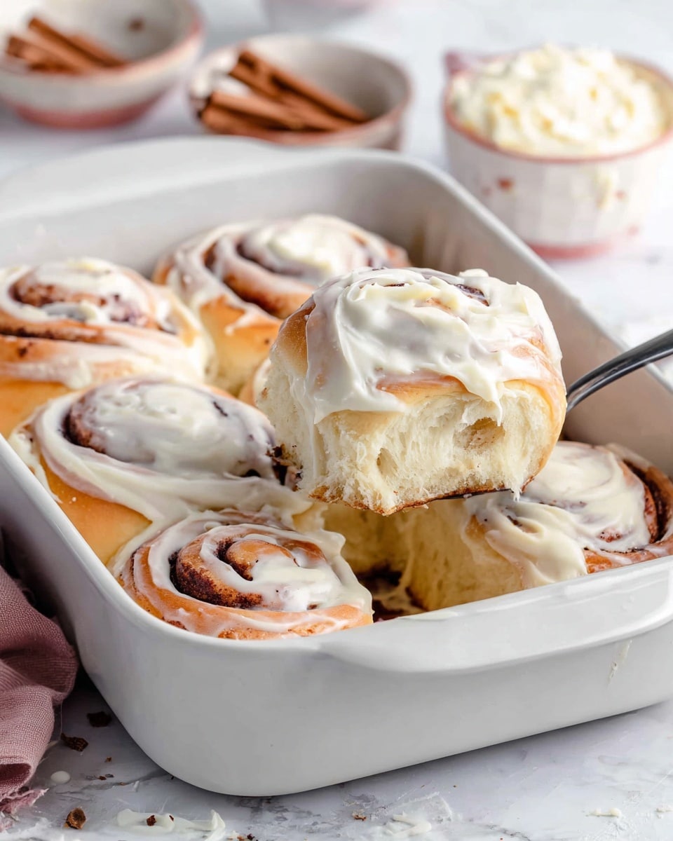The image shows six cinnamon rolls in a white rectangular baking dish on a white marbled surface, with one cinnamon roll being lifted by a spoon. Each cinnamon roll has a visible swirl layer of cinnamon brown dough and is topped with a thick, creamy white frosting that is spread unevenly, covering most of the top and sides. The rolls have a golden brown crust on top from baking, with light fluffy layers inside that look soft and airy. The background includes a small bowl of cinnamon and a wooden bowl of frosting slightly blurred. Photo taken with an iphone --ar 4:5 --v 7