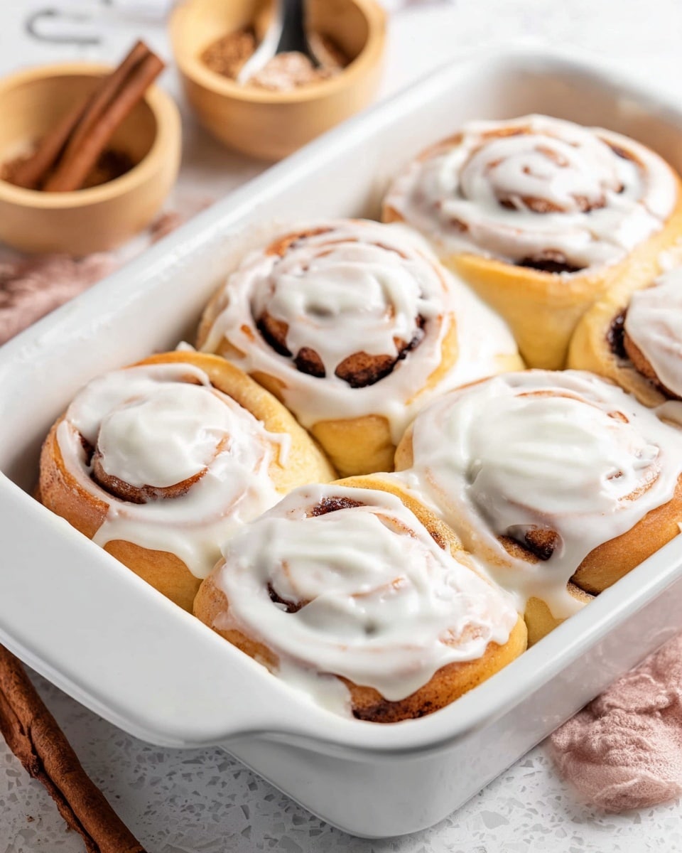 The image shows a single cinnamon roll placed in the center of a white plate on a white marbled surface. The cinnamon roll has three visible layers: a soft, light golden brown base with a slightly toasted edge, a middle swirl layer showing darker cinnamon tones mixed with warm orange-brown Highlights, and a thick, creamy white frosting generously spread on top and flowing slightly down the sides. In the background, there is a white baking dish with more cinnamon rolls and a blurred second cinnamon roll on another white plate. The lighting is bright and natural, highlighting the soft texture of the dough and the creamy frosting. photo taken with an iphone --ar 4:5 --v 7