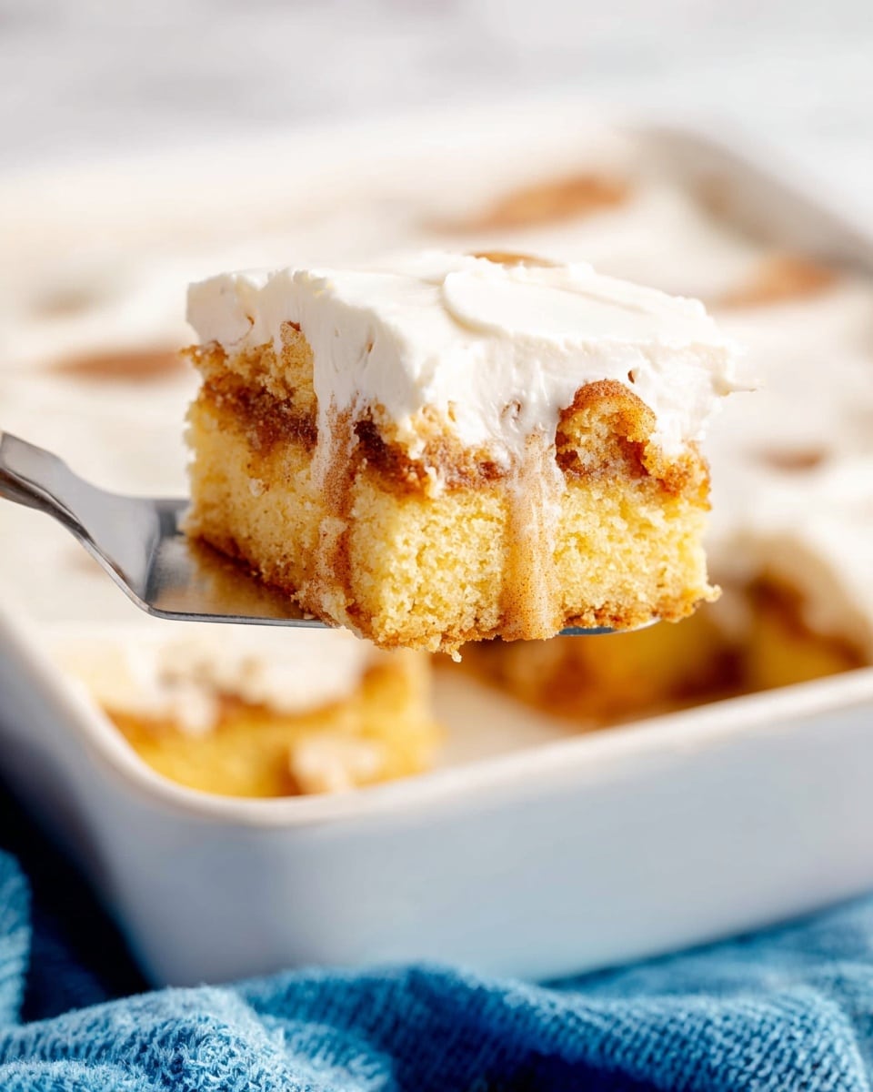 A square piece of cinnamon roll cake with three visible layers is being lifted on a spatula above a white baking dish. The bottom layer is a soft, light yellow cake base with small holes, the middle layer has swirled light brown cinnamon sugar filling mixed softly with the cake, and the top layer is a smooth, thick, white icing covering the whole piece. The white baking dish below shows more pieces covered in the same white icing, all sitting on a white marbled surface with a blue cloth nearby. photo taken with an iphone --ar 4:5 --v 7