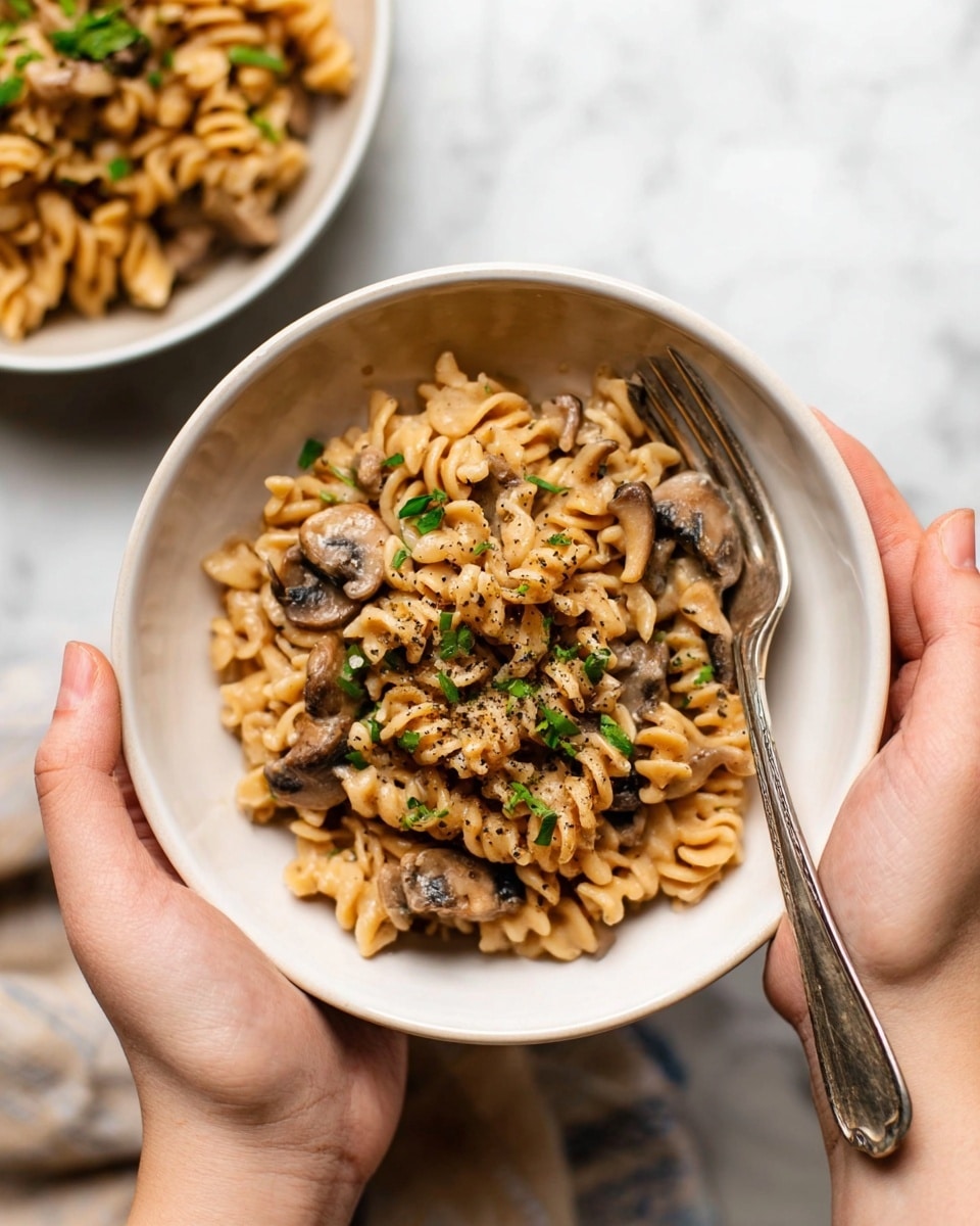 A close-up view of a white bowl filled with creamy rotini pasta mixed with sliced mushrooms in a light brown sauce. The pasta spirals are coated well, and the mushroom slices are scattered evenly throughout. A black spatula with a wooden handle is partially submerged, stirring the dish, showing small specks of black pepper on the food. The background is a white marbled texture. Photo taken with an iphone --ar 4:5 --v 7