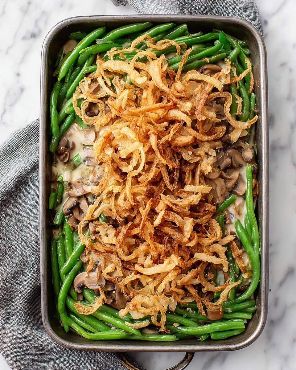 A rectangular metal baking dish filled with three visible layers: the bottom layer is bright green whole green beans that line the dish evenly; the middle layer is scattered creamy mushroom sauce with light brown sliced mushrooms mixed in; and the top layer is a thick pile of thin, golden-brown crispy fried onion strings that cover the center of the dish more heavily with some pieces spread around the edges. The dish sits on a white marbled surface with a grey cloth partially visible on the left side. Photo taken with an iphone --ar 4:5 --v 7