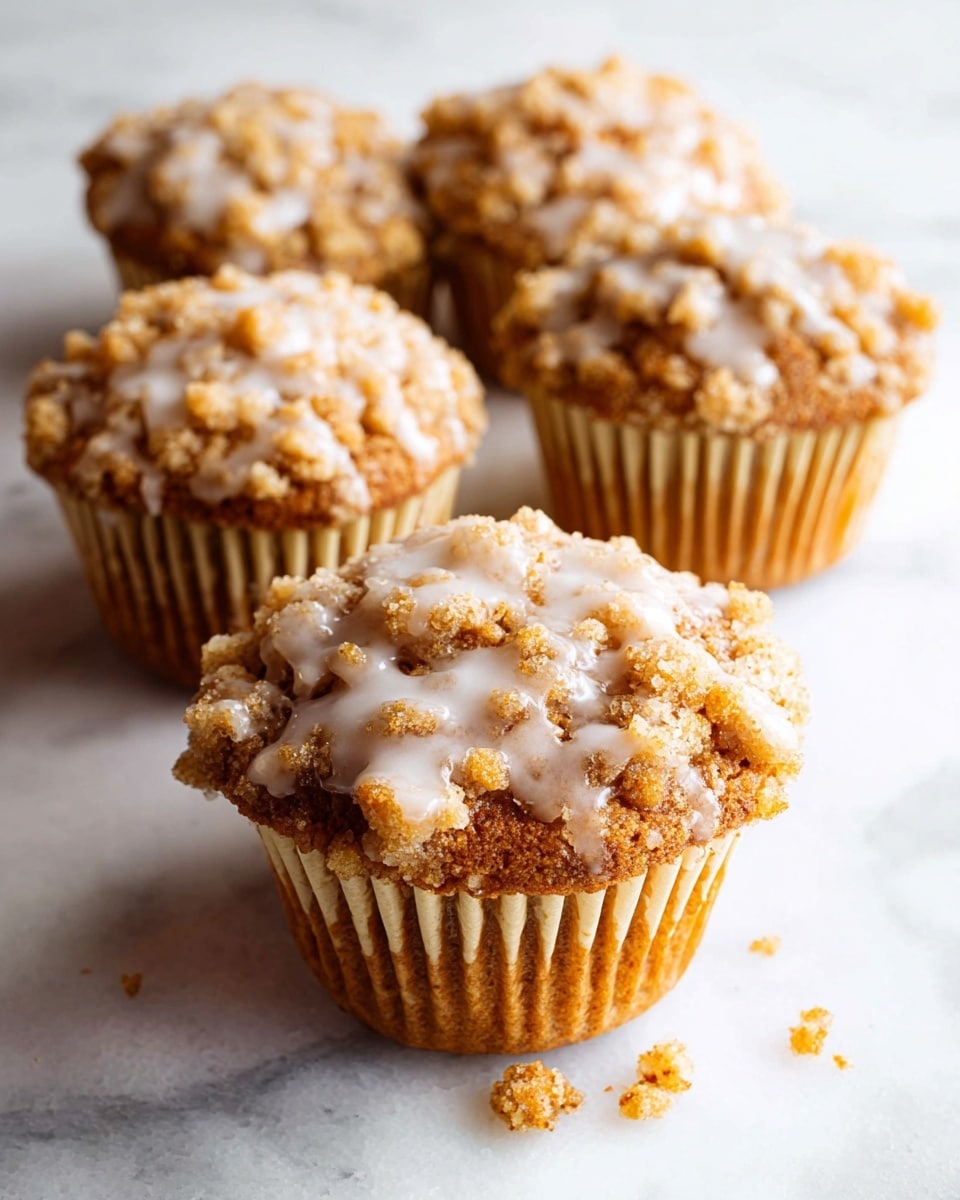 The image shows six crumb-topped muffins arranged closely on a white plate, placed on a white marbled surface. Each muffin has two main layers: a golden brown muffin base and a thick crumb topping that is light brown with a rough, crumbly texture. The crumb topping is generously drizzled with a white glaze that has a smooth, slightly shiny appearance. Some glaze has dripped onto the plate, creating small streaks and pools around the muffins. The overall look is warm and inviting, with a mix of soft and crumbly textures. photo taken with an iphone --ar 4:5 --v 7