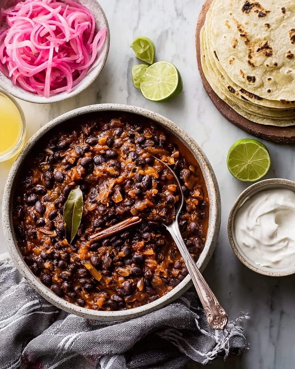 A bowl filled with a thick mixture of black beans cooked in a reddish-brown sauce with small pieces of onion and spices, topped with a visible bay leaf and a cinnamon stick, a spoon resting inside the bowl. Next to it, a white bowl containing thin slices of bright pink pickled onions. Nearby, a stack of white tortillas lying on a white marbled surface, and small lime wedges scattered around. A small white bowl with smooth white sour cream is placed with a spoon inside, and a glass with yellow liquid also sits on the surface. A gray cloth napkin with a striped pattern partially rests under the beans bowl. Photo taken with an iphone --ar 4:5 --v 7