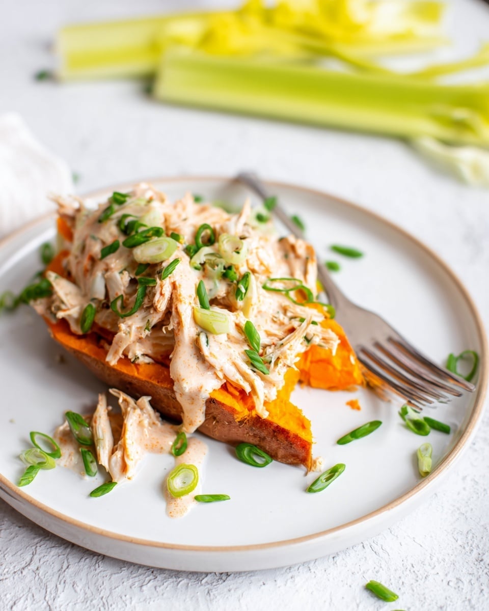 A white plate on a white marbled surface holds one thick, orange sweet potato wedge at the bottom. On top of the sweet potato is a layer of shredded light brown chicken, drizzled with a creamy white dressing and a few drops of orange sauce. Small green chive pieces are scattered over the chicken and plate. To the left side of the plate, there are a couple of pale green celery stalks. A silver fork rests on the edge of the plate. In the soft background, another plate with a similar dish is partially visible. Photo taken with an iphone --ar 4:5 --v 7