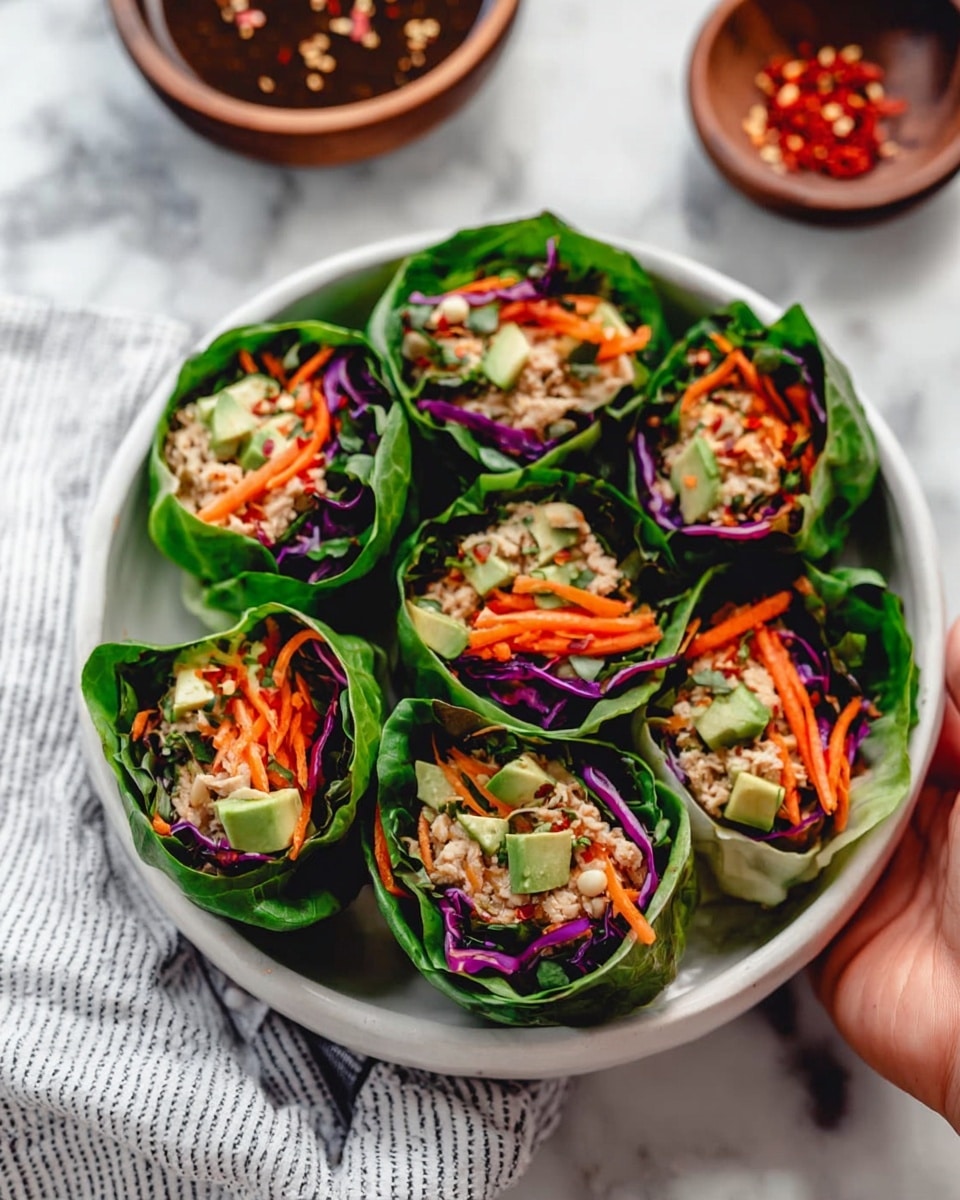 A white bowl holds six fresh green leafy wraps arranged in a circle. Each wrap has three visible layers: a dark green outer leaf, a middle layer of thinly shredded bright orange carrots and purple cabbage, and a center filled with light beige shredded filling and small pieces of pale green avocado. To the side inside the bowl, there is a small round wooden bowl filled with a dark dipping sauce sprinkled with red chili flakes. The background is a white marbled texture, and a striped cloth is partially visible near the bowl. Photo taken with an iphone --ar 4:5 --v 7