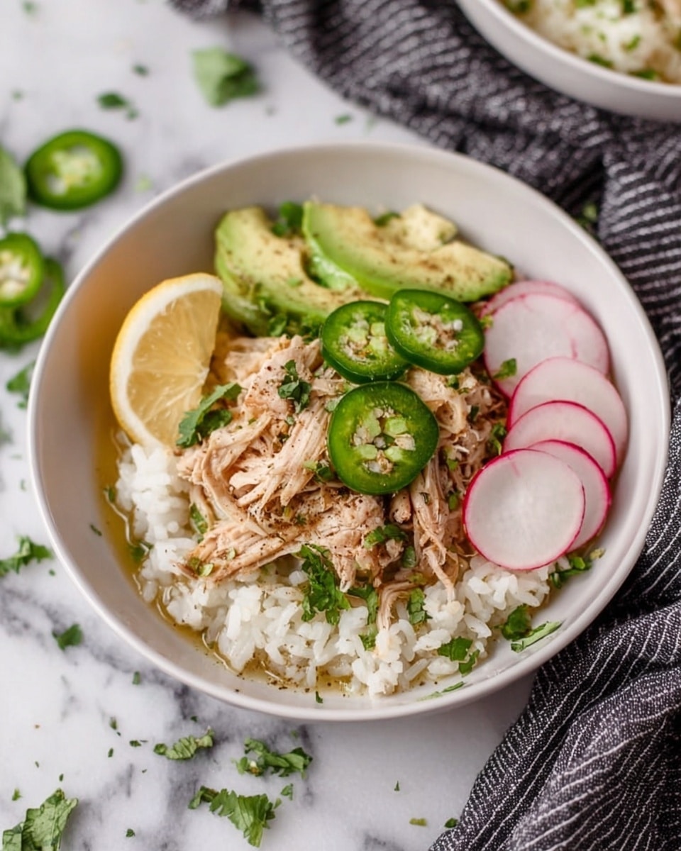 A white bowl on a white marbled surface holds a layered dish starting with a base of fluffy, white rice. On top, there is a heap of shredded light brown cooked chicken mixed with small bits of green herbs. Thin slices of bright green jalapeño peppers and pale green avocado slices are scattered over the chicken. To the side, there is a wedge of light yellow lemon and three thin, round slices of white radish with pink edges. The dish is garnished with fresh green cilantro leaves and a light sprinkle of black pepper. In the background, part of a striped black and white cloth is visible. Photo taken with an iphone --ar 4:5 --v 7