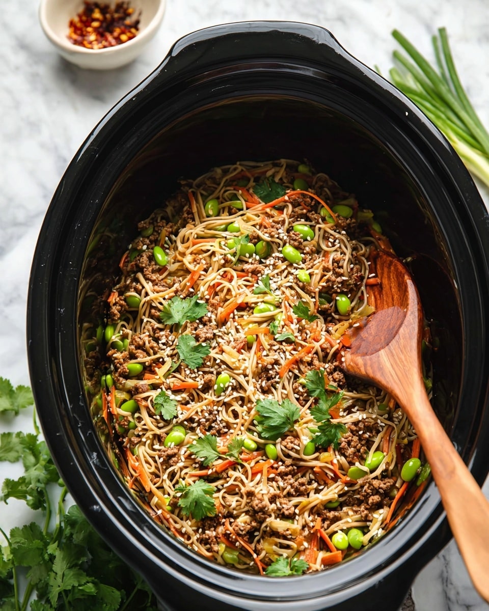 Inside a large black slow cooker, there is a mix of thin light tan noodles and small pieces of brown cooked meat layered with thin orange carrot strips and bright green edamame beans. The dish is topped with fresh green cilantro leaves and sliced green onions scattered around. A light sprinkling of white sesame seeds adds texture, and a wooden spoon is resting inside the cooker, partly buried in the food. The background is a white marbled surface with some scattered small dishes of red pepper flakes and extra green onions. photo taken with an iphone --ar 4:5 --v 7