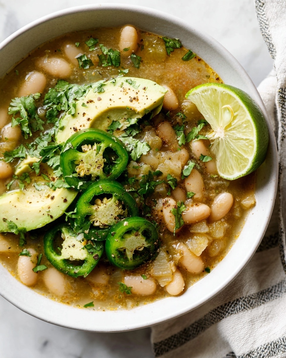 A white bowl filled with a soup consisting of creamy beige beans and soft yellow chunks of cooked vegetables in a light broth, topped with bright green slices of fresh avocado and dark green rings of jalapeño pepper showing seeds inside, sprinkled with chopped cilantro leaves and fresh black pepper. A wedge of lemon, pale yellow with a slightly translucent texture, rests on the edge of the bowl. The bowl sits on a white marbled surface with a striped cloth nearby. Photo taken with an iphone --ar 4:5 --v 7