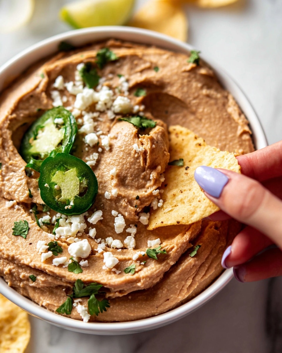 A close-up image of a white plate filled with a smooth brown layer of refried beans spread evenly across the surface. On top of the beans are small white crumbles of cheese scattered around and bright green slices of jalapeño with fresh cilantro leaves placed for garnish. A woman's hand with pale lavender nail polish is dipping a light yellow, slightly crisp tortilla chip into the beans. The scene is set on a white marbled textured surface, highlighting the warm and fresh colors of the dish photo taken with an iphone --ar 4:5 --v 7