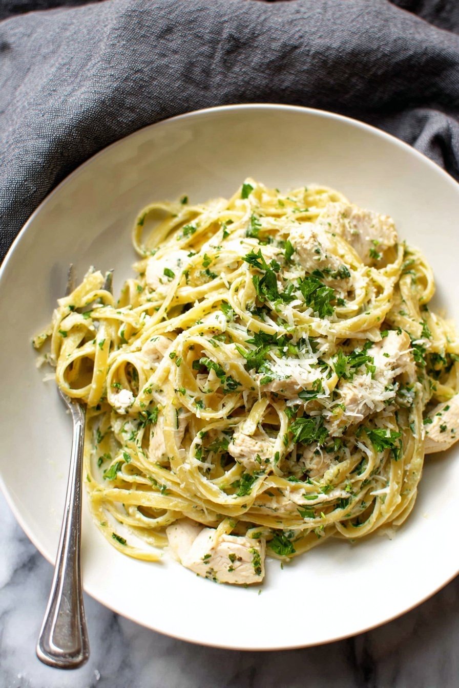 A white bowl filled with pasta, with two layers visible: the bottom layer is light yellow fettuccine noodles, and the top layer has small white pieces of chicken mixed with bright green chopped herbs, all coated in a creamy sauce; a silver fork rests inside the bowl, and the bowl is set on a soft gray fabric over a white marbled surface photo taken with an iphone --ar 4:5 --v 7