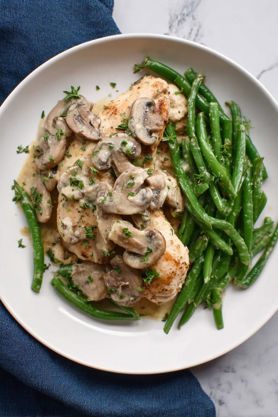 A white round plate holds a dish with three main layers: the bottom layer is green beans with a fresh, slightly glossy texture, arranged loosely across the plate. On top of the green beans is a creamy mushroom sauce with sliced mushrooms, light tan in color with some darker edges, spread over and slightly around the middle. The top layer features two small pieces of cooked light brown meat resting under and near the mushroom sauce, with fresh green herbs sprinkled over everything. The plate sits on a white marbled surface, and a dark blue cloth is partly visible near the edge. photo taken with an iphone --ar 4:5 --v 7